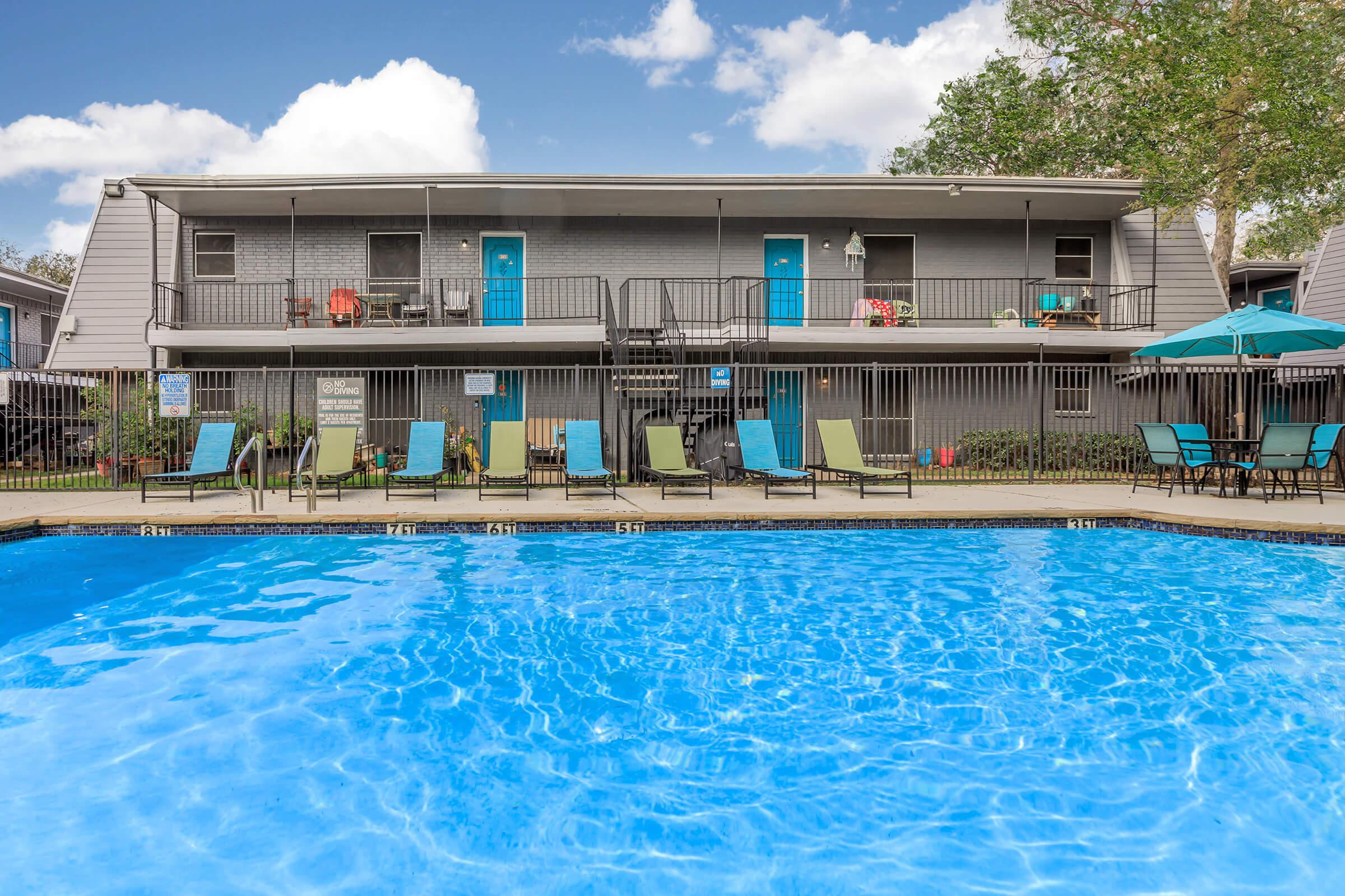 A clear blue swimming pool in the foreground, with several lounge chairs arranged around it. In the background, a two-story building features light gray siding, bright blue doors, and a balcony with chairs. Lush greenery surrounds the area, and a few umbrellas provide shade near the poolside.