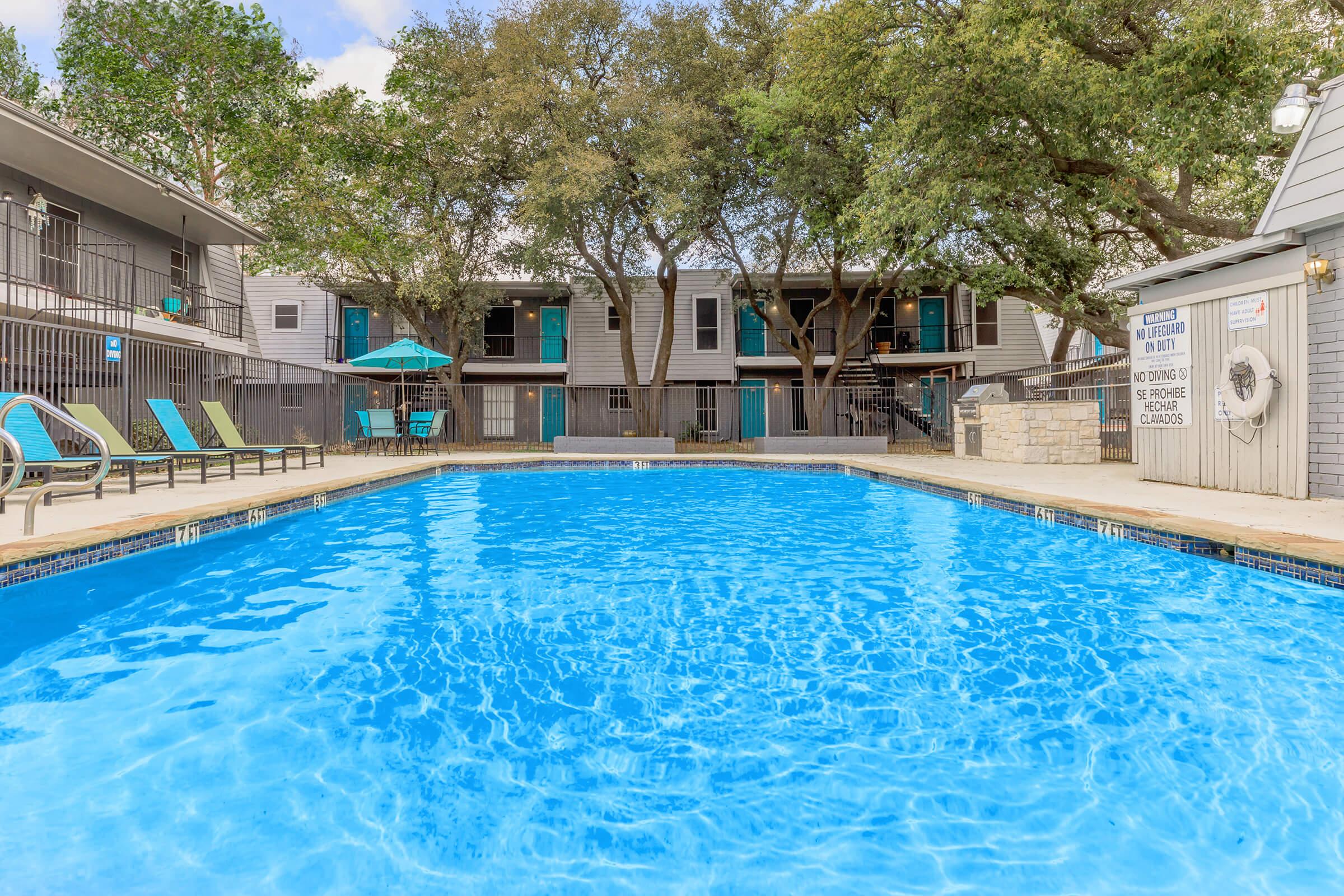 A clear blue swimming pool surrounded by lounge chairs. In the background, there are two-story buildings with teal doors, shaded by large trees. The setting is calm and inviting, ideal for relaxation, with a few umbrellas providing shade.