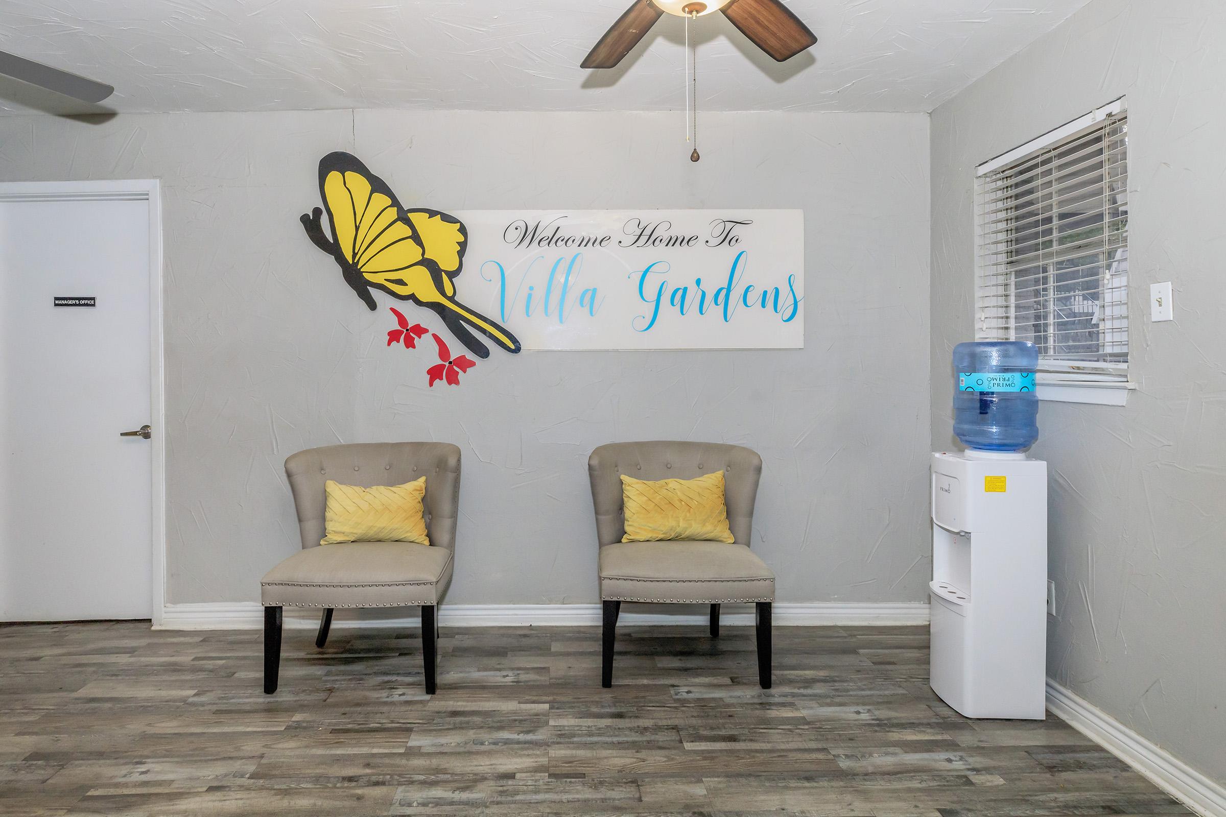 A welcoming interior space featuring two gray chairs with yellow cushions, a water cooler, and a large sign that reads "Welcome Home To Villa Gardens." A butterfly illustration is depicted next to the text, enhancing the cozy and inviting atmosphere.