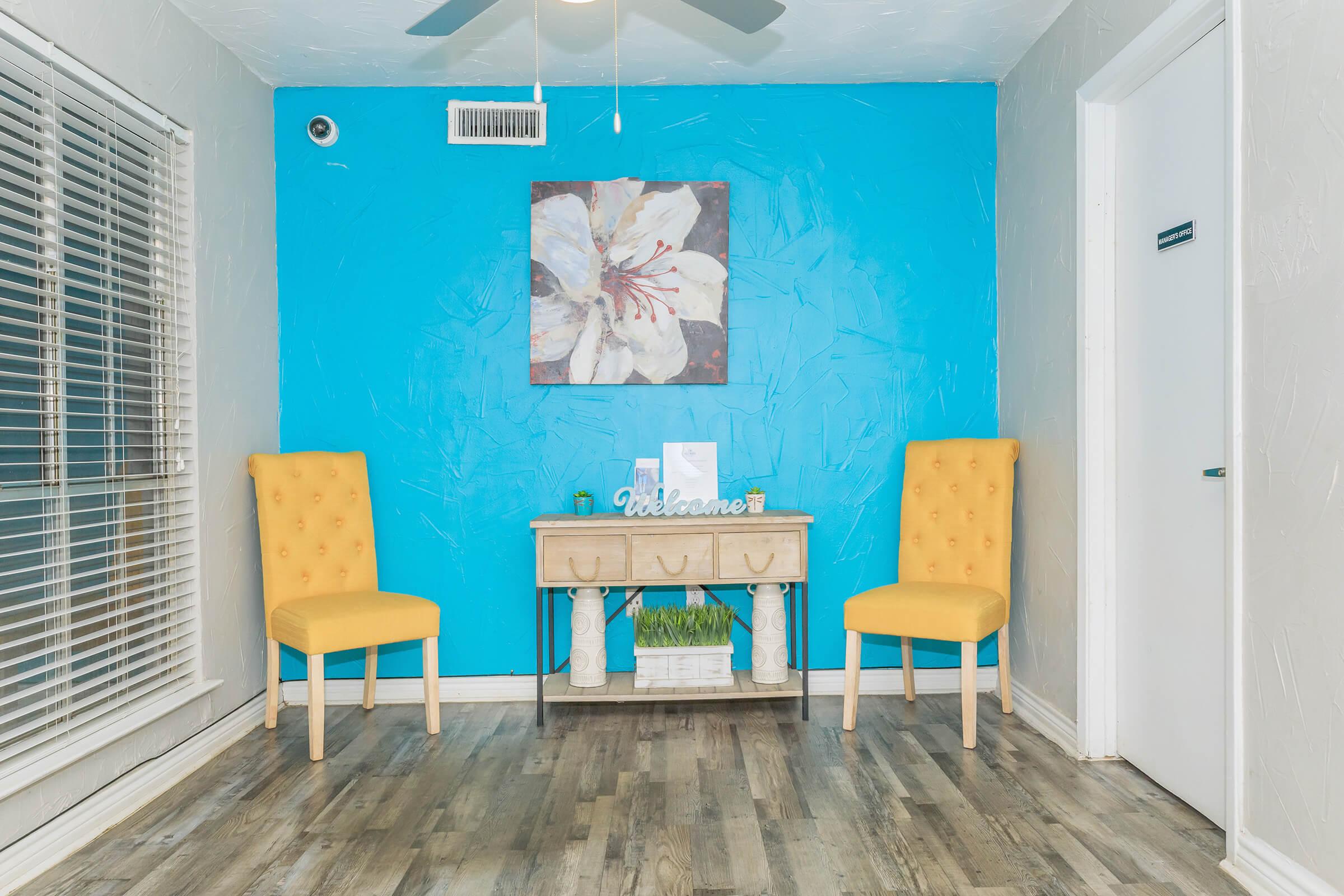 A bright interior featuring a turquoise accent wall with a floral painting, two yellow upholstered chairs, and a small wooden table with decorative items. The floor is wood-like, and there are natural light sources from the window and ceiling fan. A welcoming atmosphere is created in this cozy space.