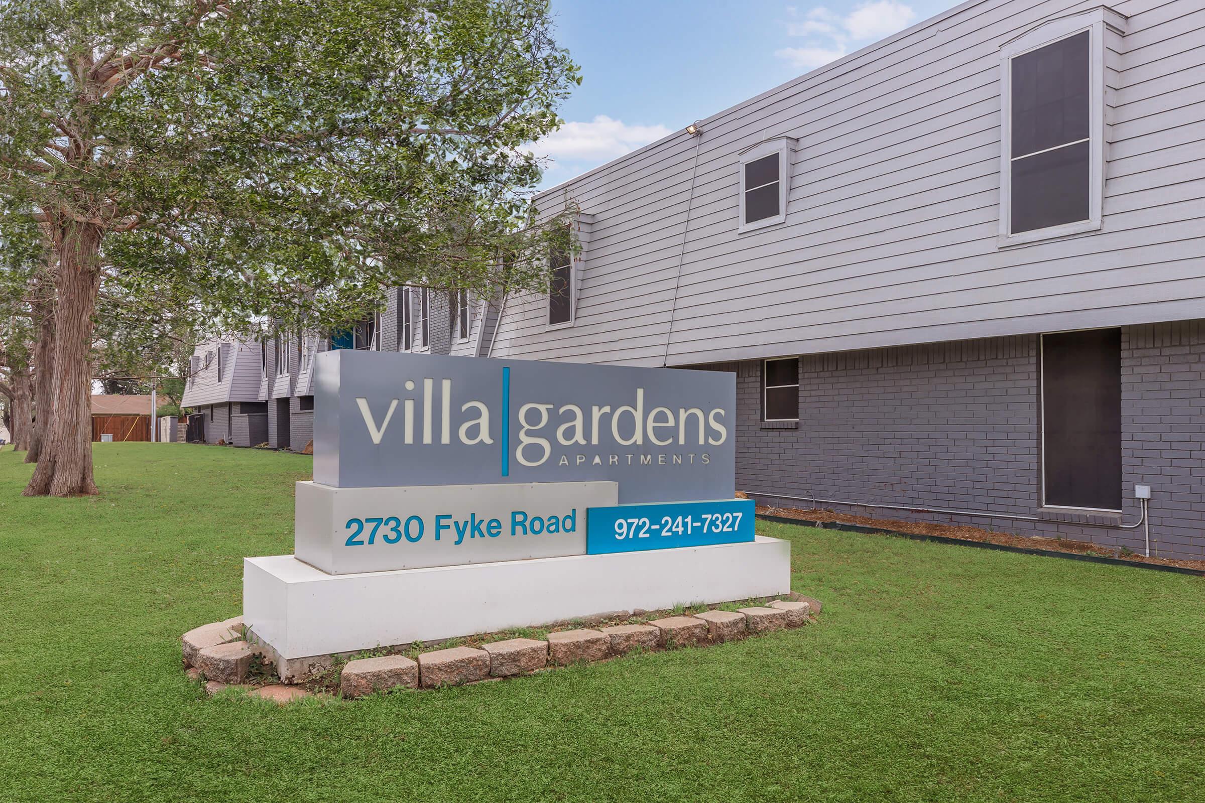 Sign for Villa Gardens Apartments located at 2730 Fyke Road, featuring the name "Villa Gardens Apartments" in bold letters. The sign includes a contact number, 972-247-7327, and is situated in a green lawn area with modern apartment buildings in the background.