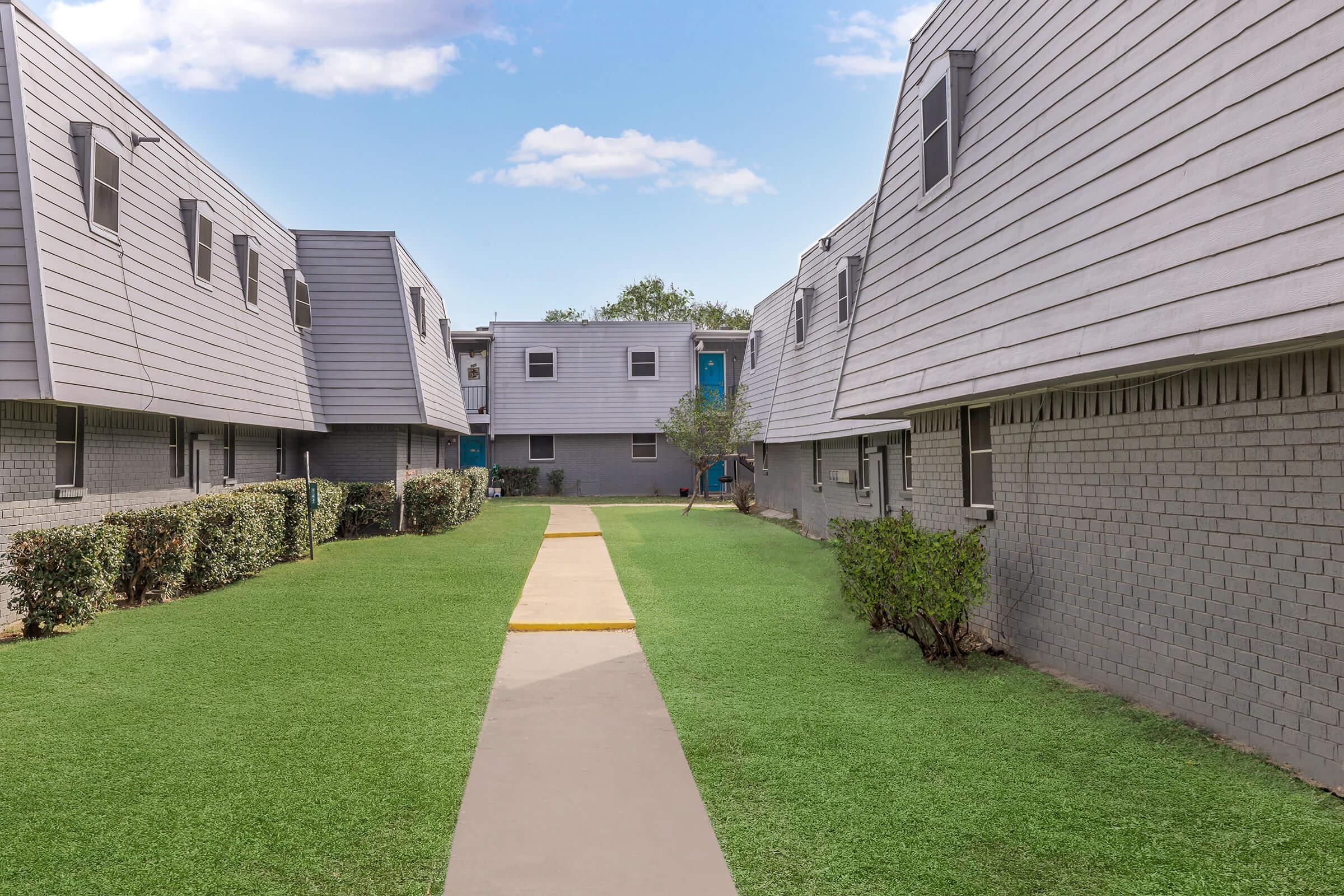 A well-maintained courtyard surrounded by two-story apartment buildings with a landscaped grassy area. A paved path runs through the center, leading deeper into the complex. The buildings feature gray siding and smaller windows, creating a peaceful residential atmosphere. Blue doors add a pop of color to the setting.