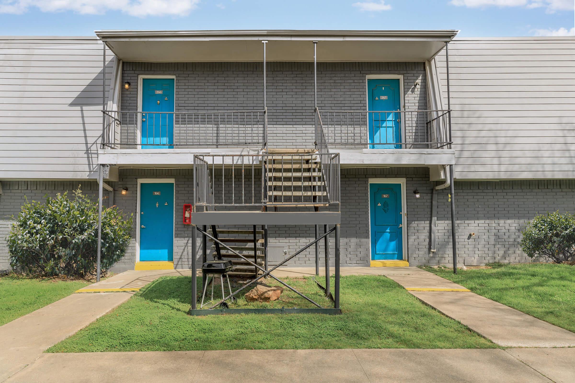 Two-story apartment building with gray brick exterior and bright blue doors. A set of metal stairs leads to the upper level. Green grass surrounds the entrance, and a fire extinguisher is visible near the bottom of the stairs. The sky is partly cloudy, creating a bright, inviting atmosphere.