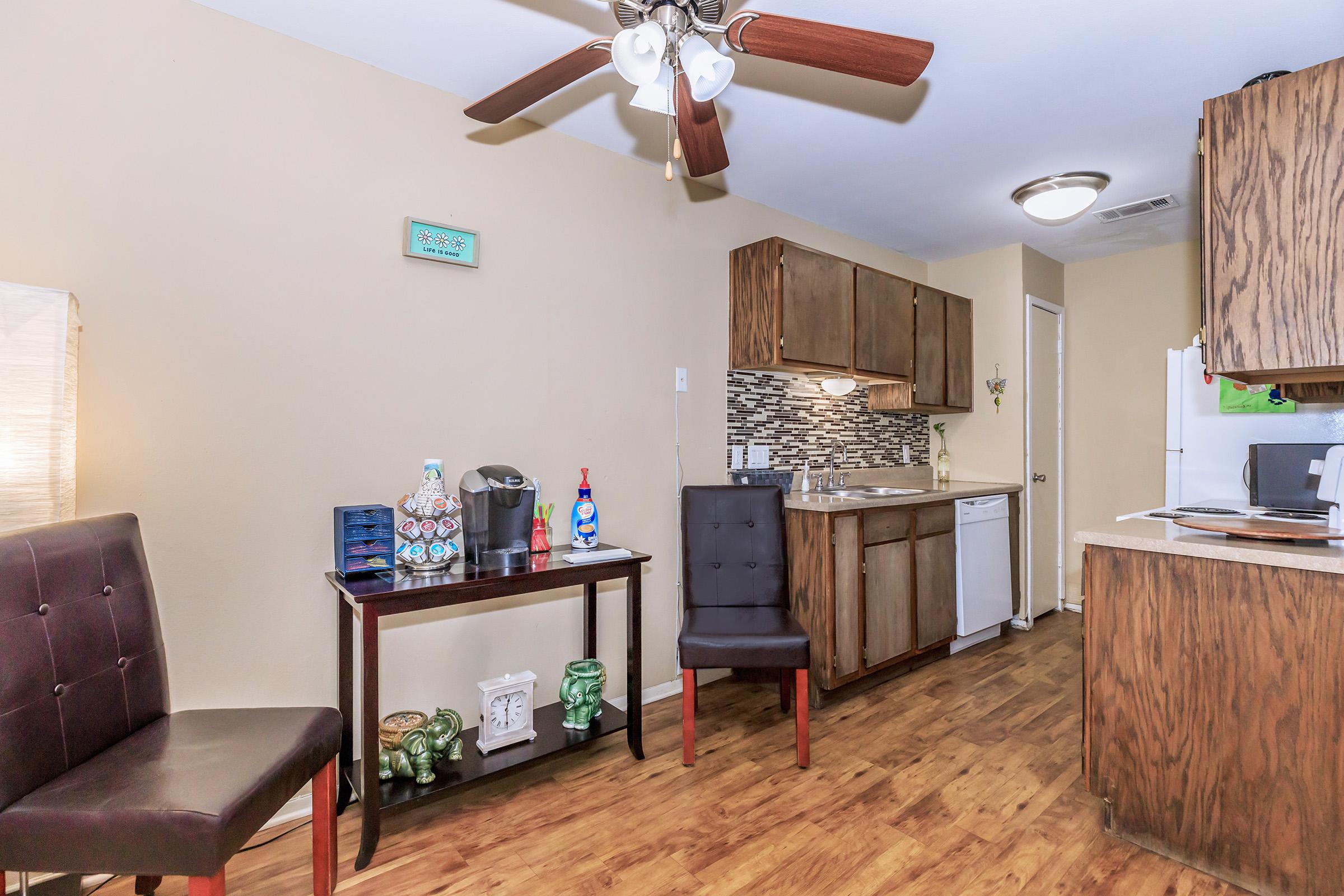 A small kitchen area featuring dark wood cabinetry, a modern backsplash, and essential appliances. There's a dining table with two chairs and a decorative table in the foreground. The floor is wood-like, and the walls are a light beige color. A ceiling fan is visible above.