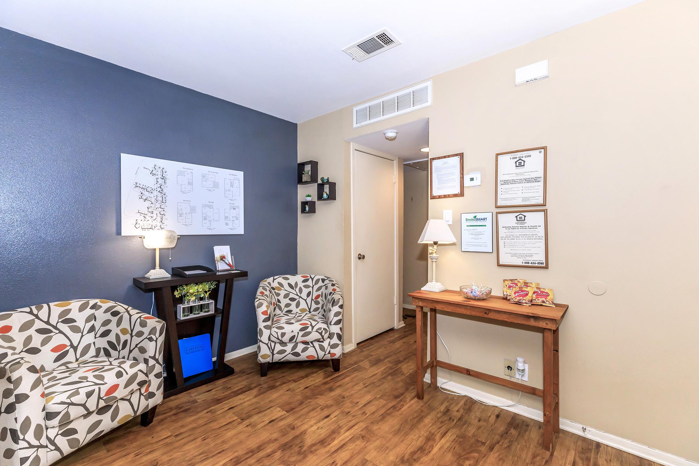 A cozy waiting room featuring two patterned armchairs, a small table with a lamp, and a wooden table displaying informational documents and snacks. The walls are painted in a mix of light and dark colors, creating a welcoming atmosphere. A door leads to another room, enhancing the inviting space.
