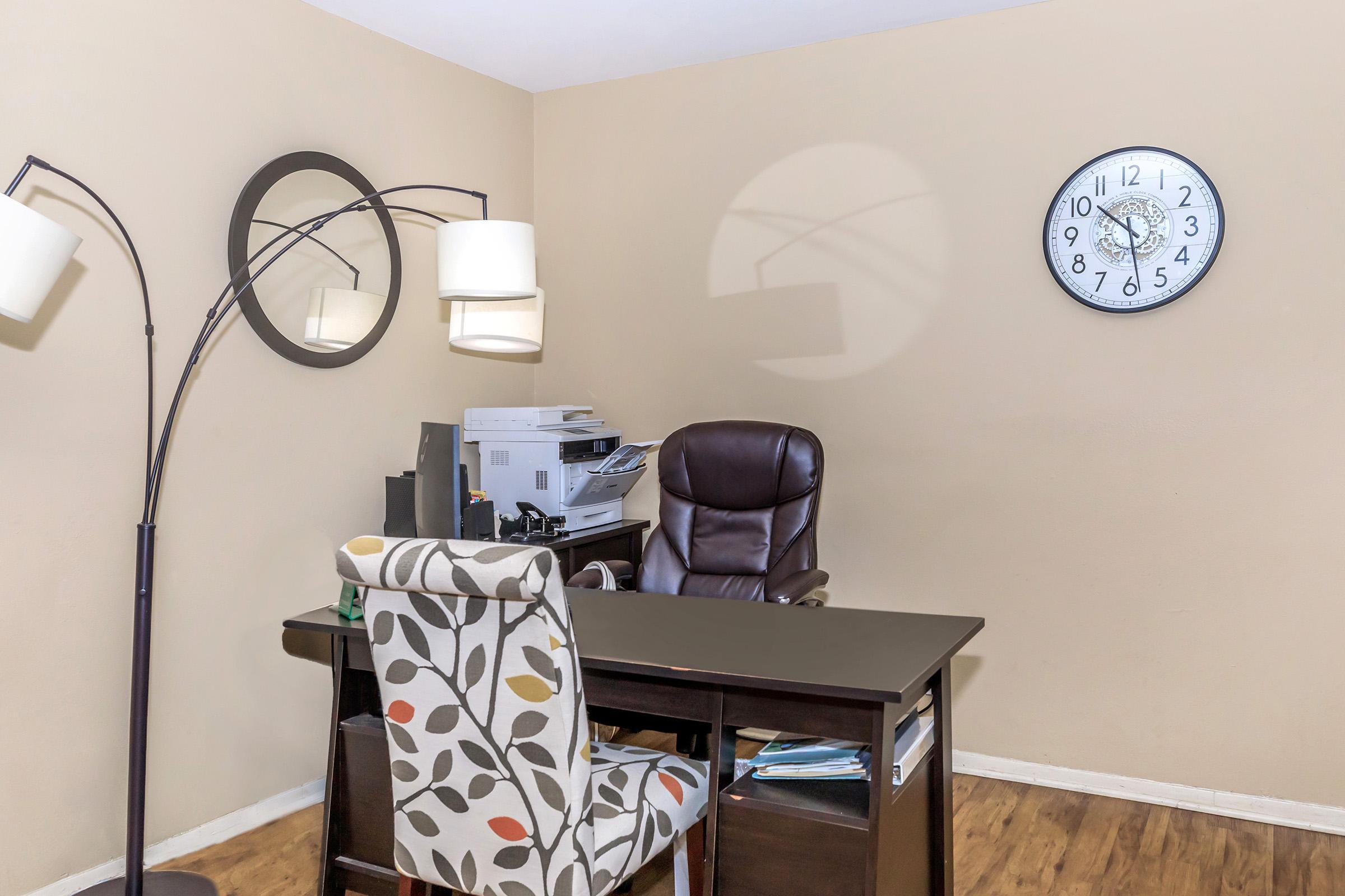 An office space featuring a dark wooden desk with a chair, a printer, and a modern floor lamp. A decorative chair with a leaf pattern is positioned nearby. A wall clock and two circular mirrors are mounted on the beige wall, creating a cozy and functional atmosphere. The floor has a warm wooden finish.