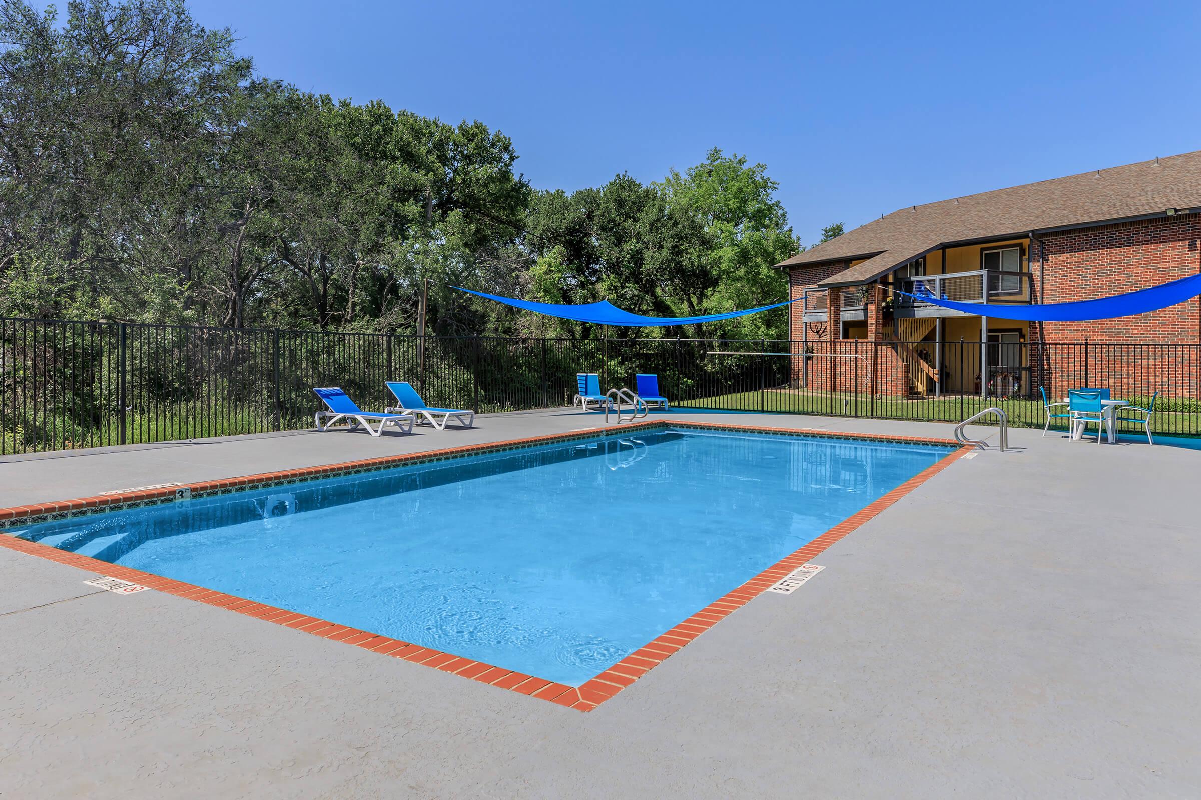 A clear blue swimming pool surrounded by lounge chairs and shaded by blue canopies. In the background, there are trees and a brick building with balconies, set under a clear blue sky. The pool area is well-maintained and inviting for relaxation.
