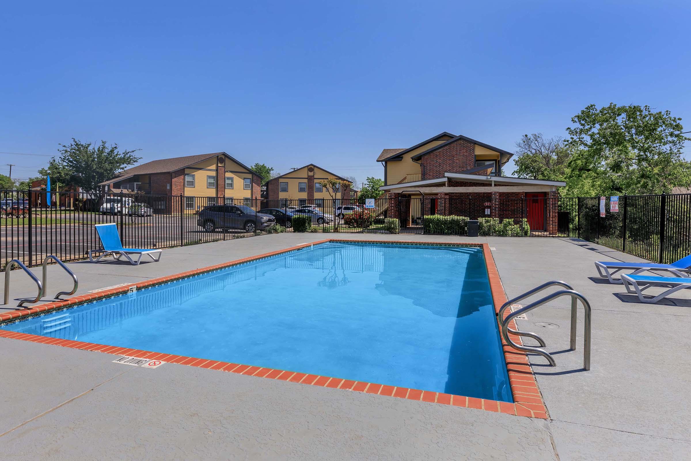 A clear blue swimming pool surrounded by a concrete deck, lounge chairs, and a fenced area. In the background, there are two multistory residential buildings and trees under a clear blue sky. The scene is bright and inviting, perfect for relaxation and leisure.