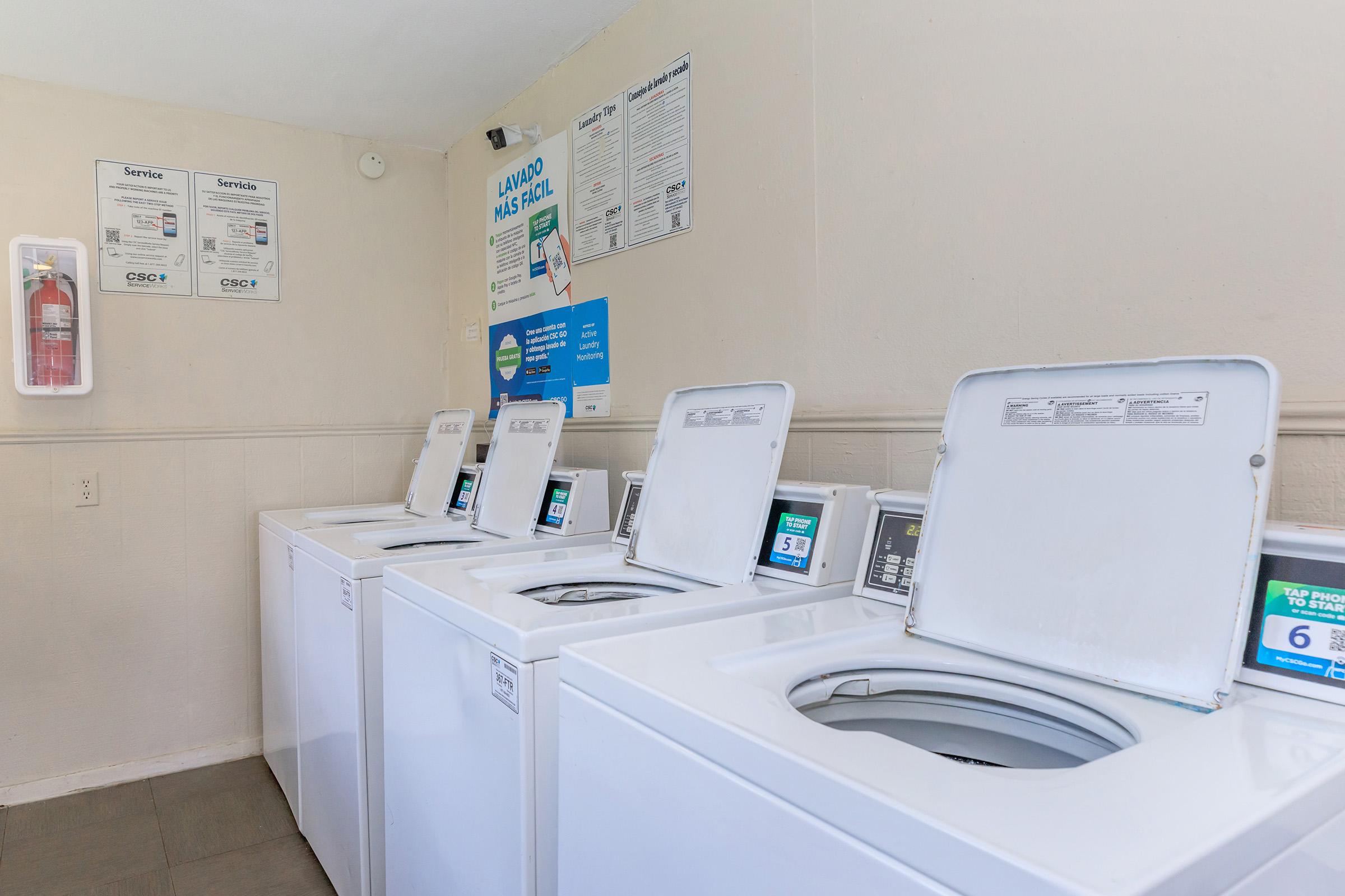 A clean and well-lit laundromat featuring several white washing machines lined up in a row. Each machine has a digital display. The walls are painted light colors, and there are informational posters about laundry services on the wall. A fire extinguisher is also visible on the left.