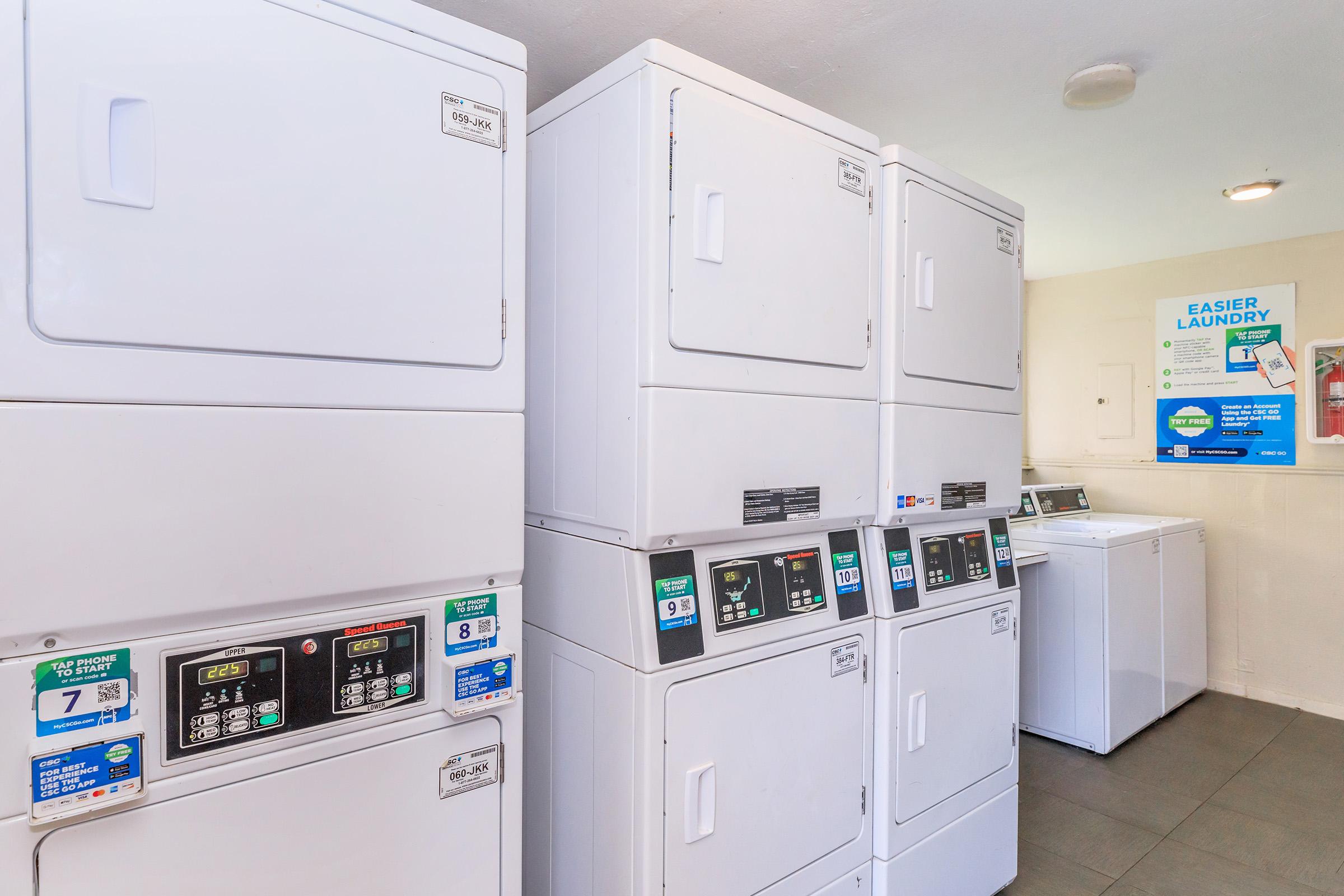 A modern laundry room featuring stacked white washer and dryer units, along with a row of standard washing machines. Various control panels and displays are visible, indicating settings and instructions for operation. A poster on the wall highlights laundry tips. The space is brightly lit and well-organized.
