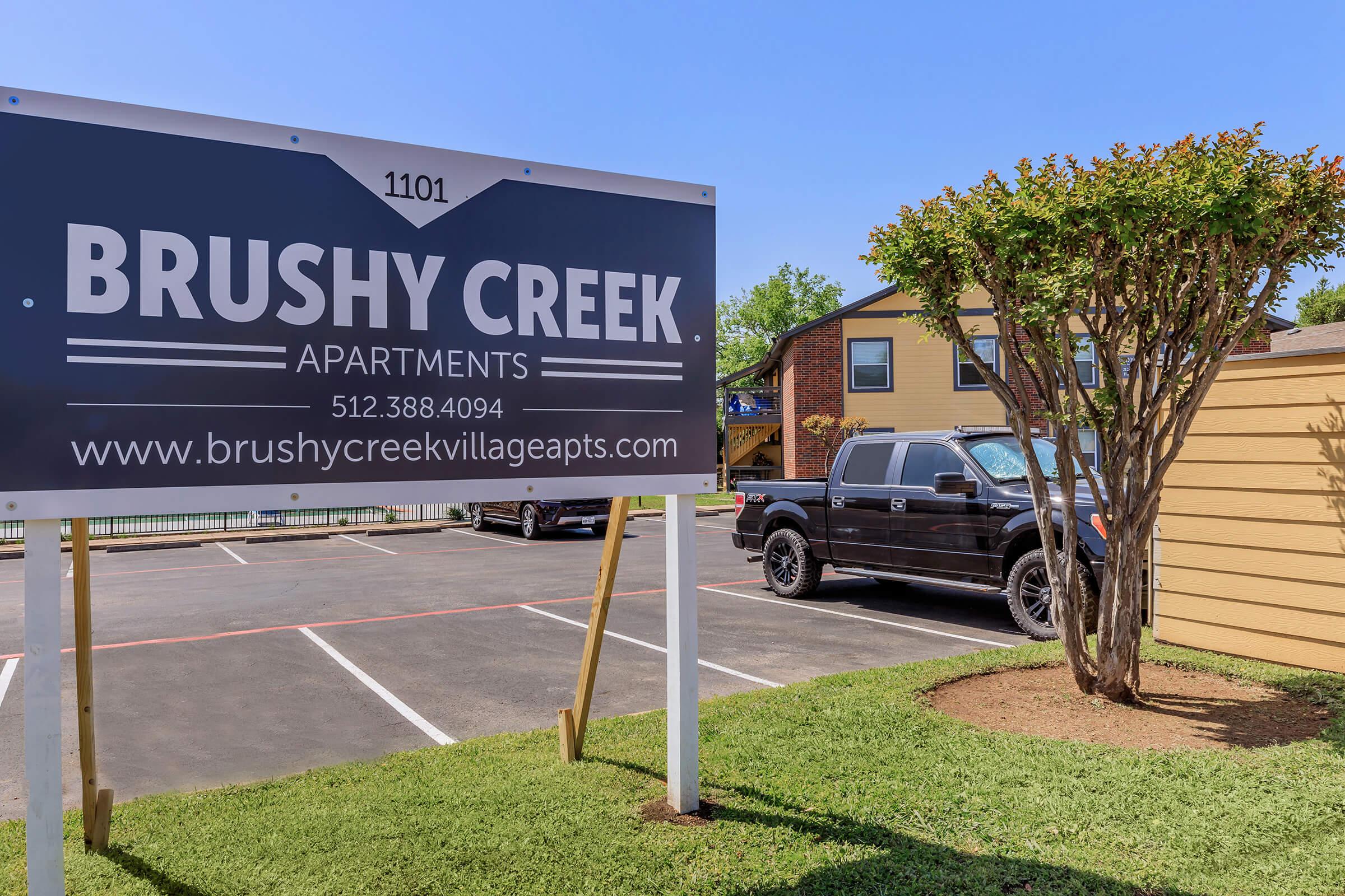 Sign for Brushy Creek Apartments at 1101, featuring the contact number 512-388-4094 and website. The foreground shows a black truck parked near the sign in a well-maintained area with grass and trees, while apartment buildings are visible in the background.