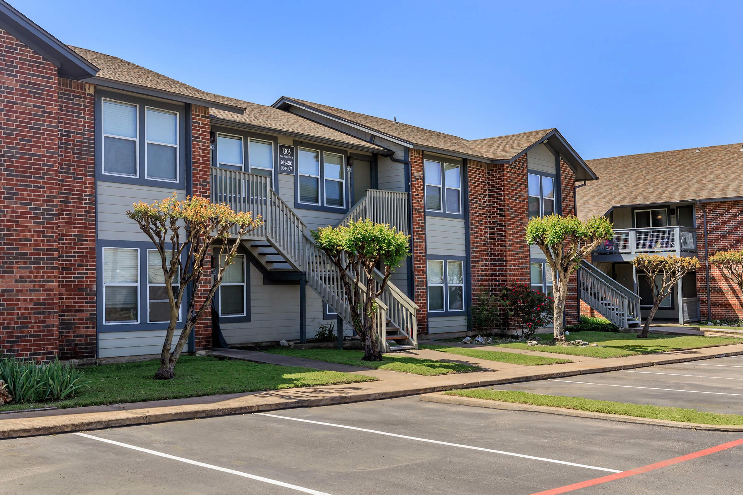 A view of a multi-unit apartment building featuring brick exteriors and wooden staircases. The landscaped area includes small trees and grass. Parking spaces are visible in front of the building on a sunny day.