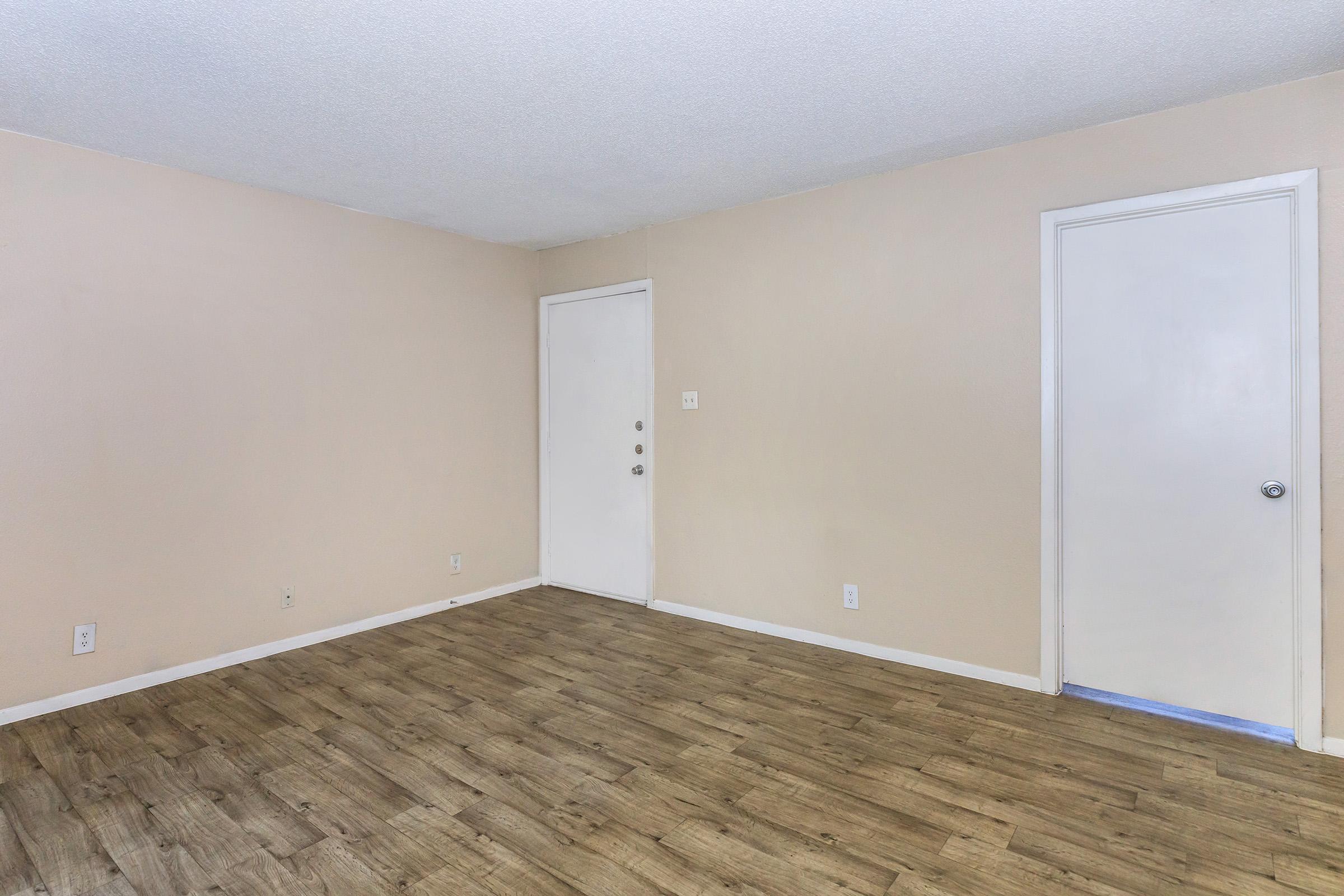 Empty room with light beige walls and a wooden floor. There is a closed white door on one wall and another door leading outside on an adjacent wall. The space is well-lit, conveying a clean and simple aesthetic, suitable for various uses or furnishings.