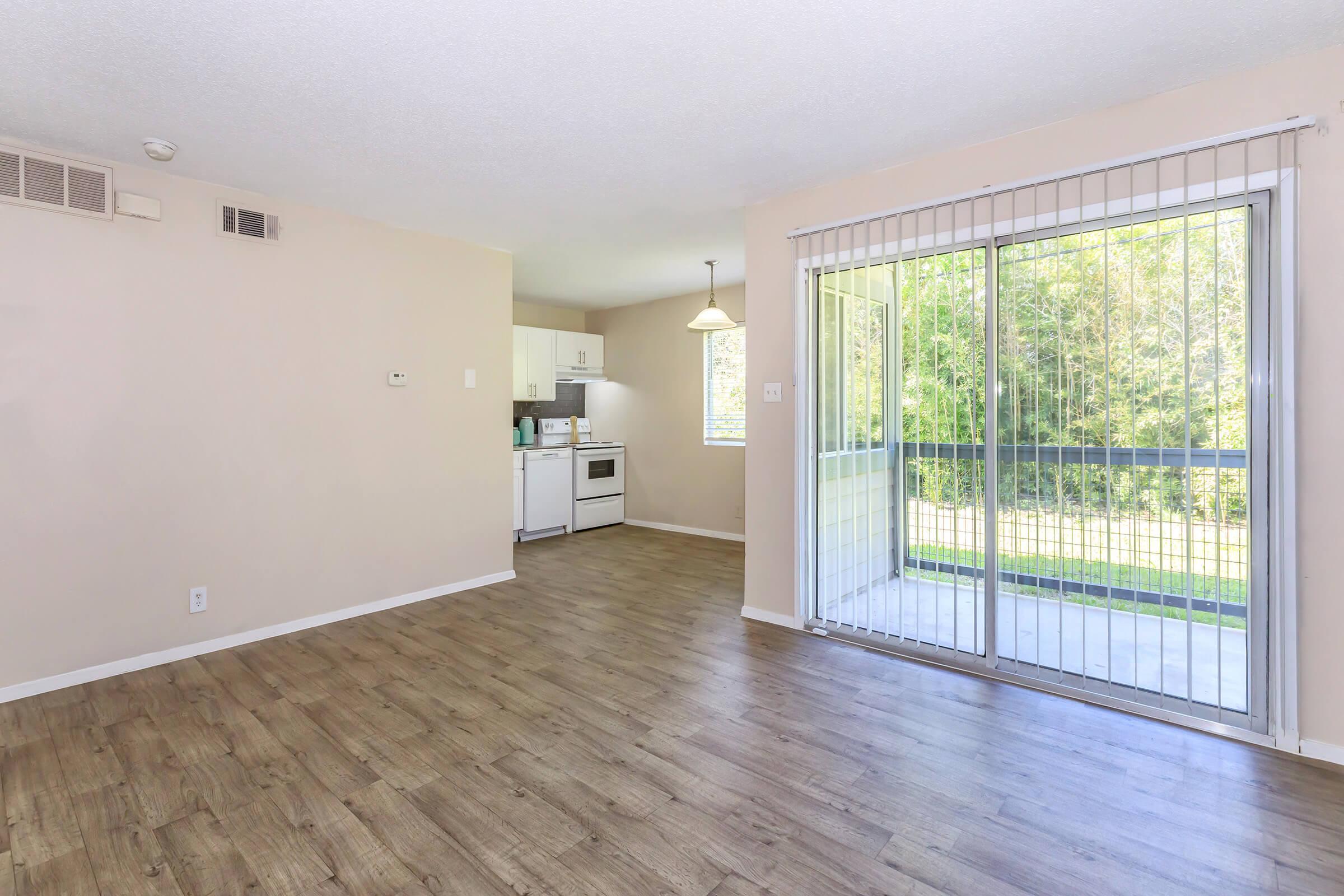 Spacious living area featuring laminate flooring, natural light, and a sliding glass door leading to a patio. The kitchen area is visible in the background, equipped with white appliances and a small dining space. The walls are painted in a soft, neutral color, enhancing the bright atmosphere.