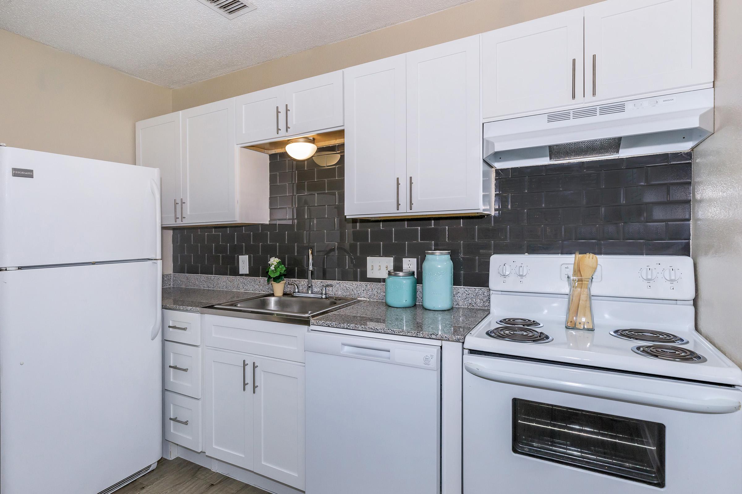 A modern kitchen featuring white cabinets, a gray countertop, and stainless steel appliances. The space includes a refrigerator, dishwasher, and stove, with a black tile backsplash and blue decorative vases. A small plant adds a touch of greenery to the design.