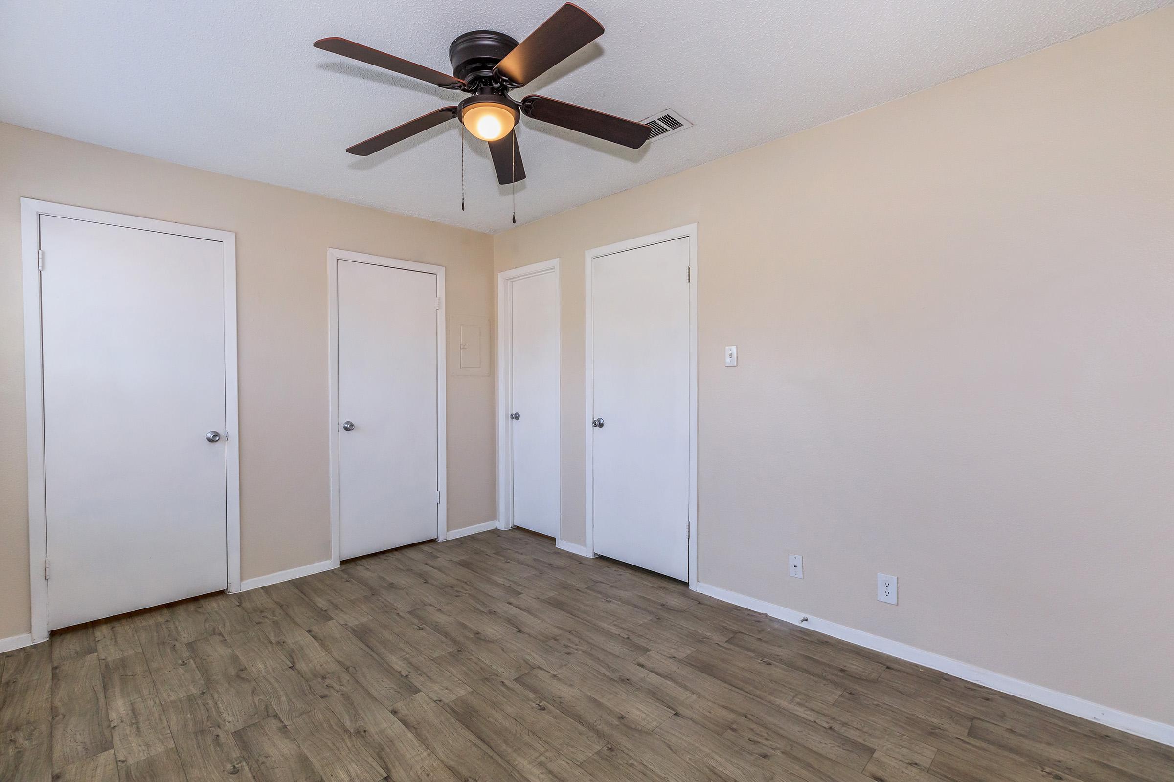 A neutral-toned room featuring a ceiling fan, four doors on the walls, and light brown wood-like flooring. The space appears clean and unfurnished, ideal for a variety of uses.