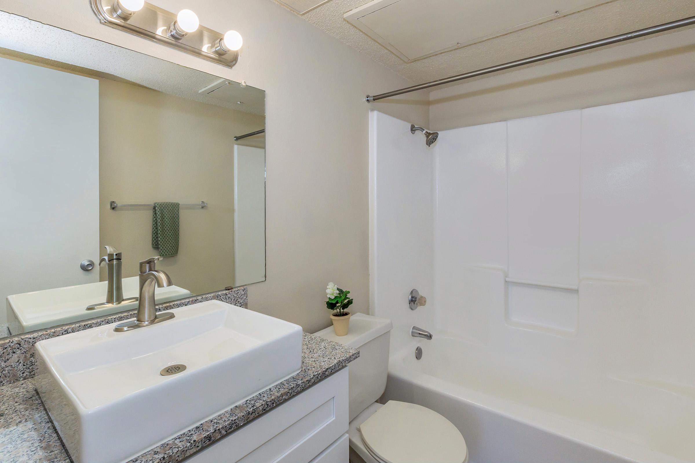 Modern bathroom featuring a white sink with a sleek faucet, a large mirror above, and a white bathtub with a shower. The wall has a neutral color, and a small potted plant adds a touch of decor. A hand towel is hanging on a rack nearby, and the overall setting is clean and minimalistic.