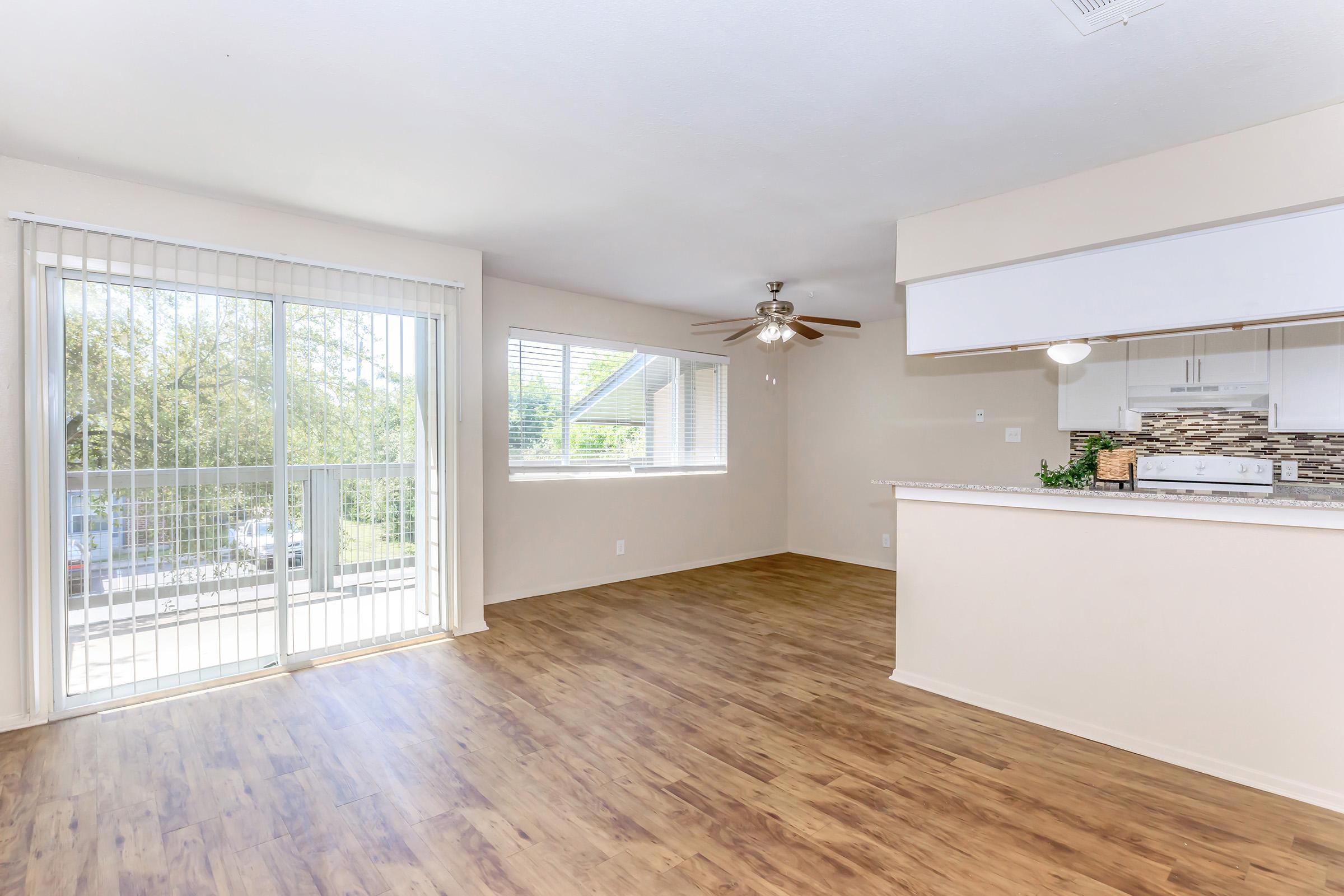 A bright, spacious living area with wood-style flooring, featuring a ceiling fan, large windows with blinds, and an open kitchen with a light-colored countertop. Natural light fills the room, creating a welcoming atmosphere. The view includes greenery outside through the sliding glass door.