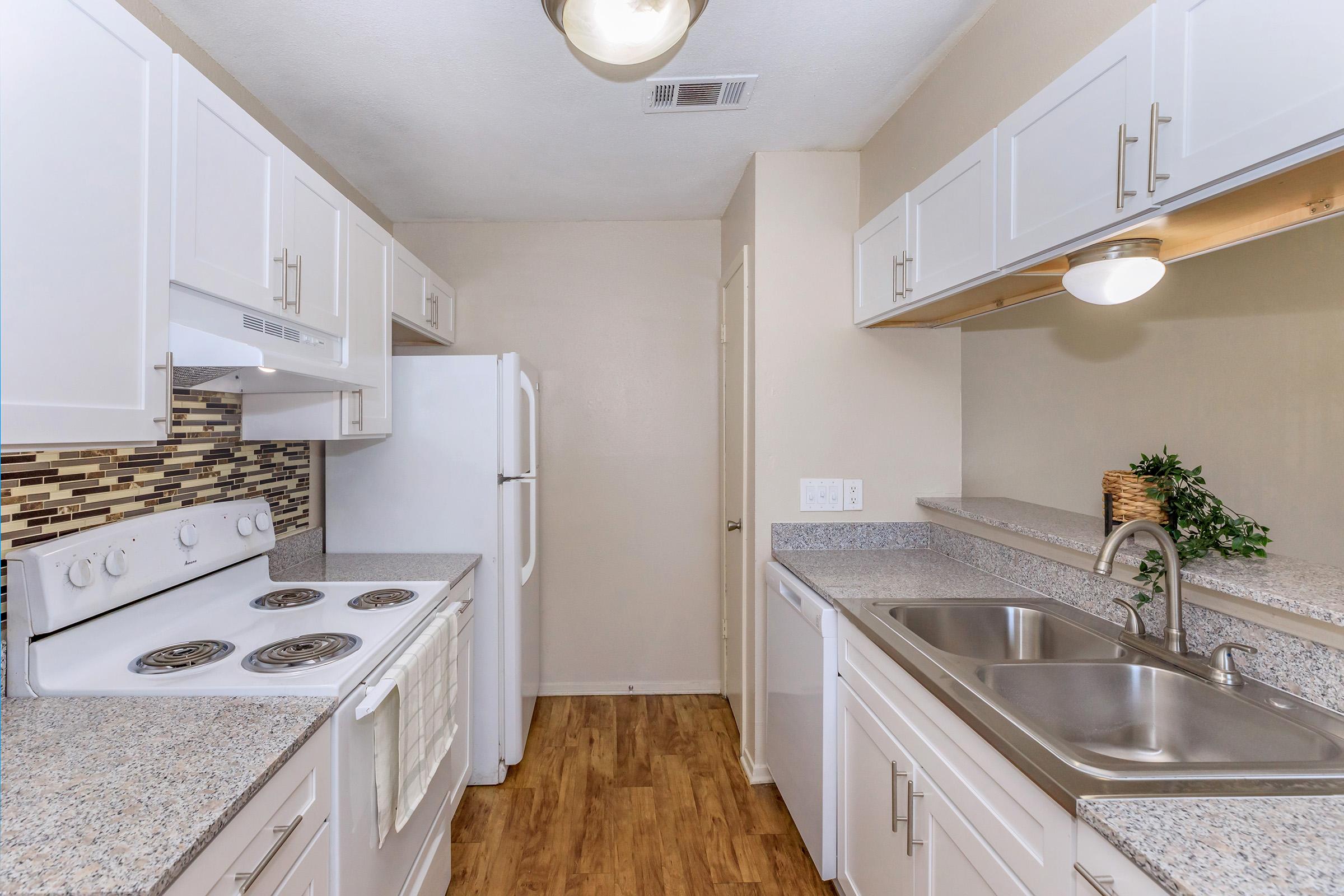 Modern kitchen with white cabinetry, granite countertops, and a light-colored backsplash. The space includes a refrigerator, an electric stove, and a double sink. Wood-style flooring adds warmth, while overhead lighting brightens the area.