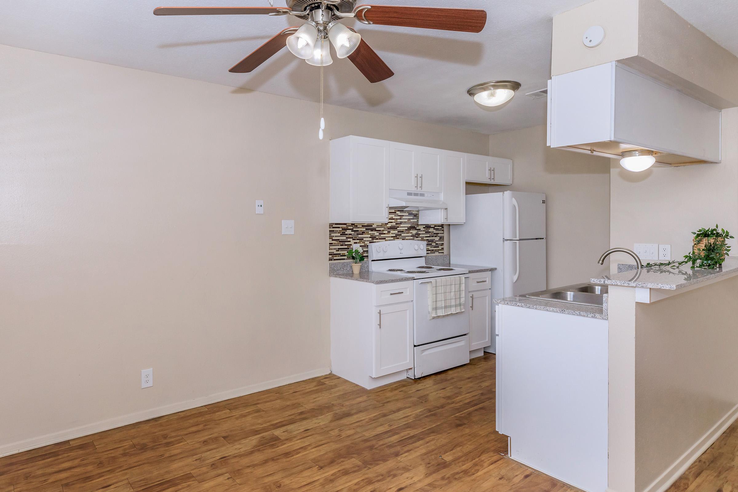 A modern kitchen featuring white cabinets, a stainless steel refrigerator, and a stove. The countertop is gray with a small sink and a potted plant. A light fixture hangs from the ceiling, and wooden flooring adds warmth to the space. The walls are painted a soft beige, creating a bright, inviting atmosphere.