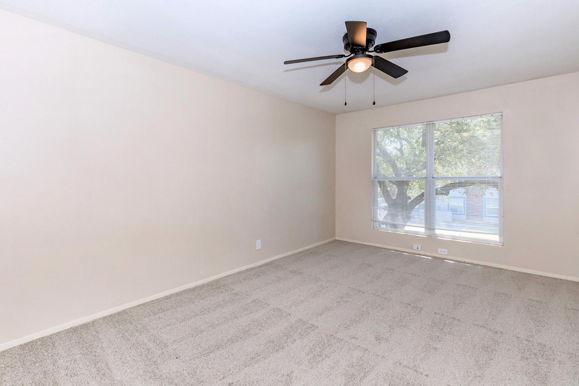 A spacious, empty room with light beige walls and a light-colored carpet. It features a ceiling fan with five blades and a large window providing natural light, framed by clear glass. The overall atmosphere is bright and open, ideal for various uses or decoration.