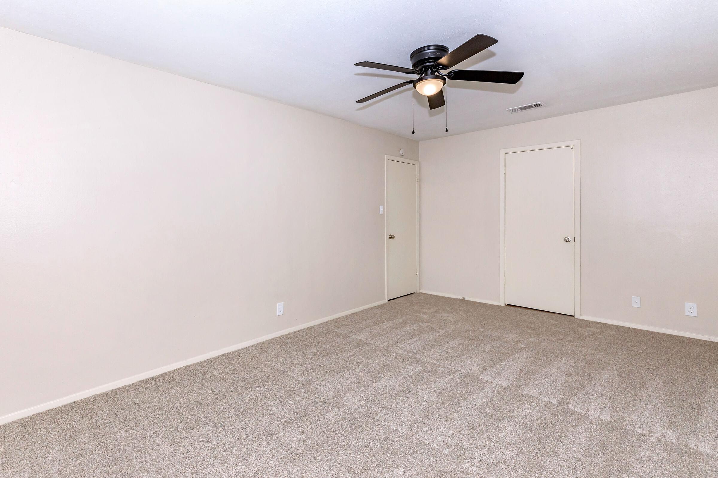 A spacious, empty room with light beige walls and plush carpet. A ceiling fan with dark blades is mounted in the center. There is a closed door on the far wall and a second door on the right, both matching the wall color. Natural light is absent, creating a neutral and inviting atmosphere.