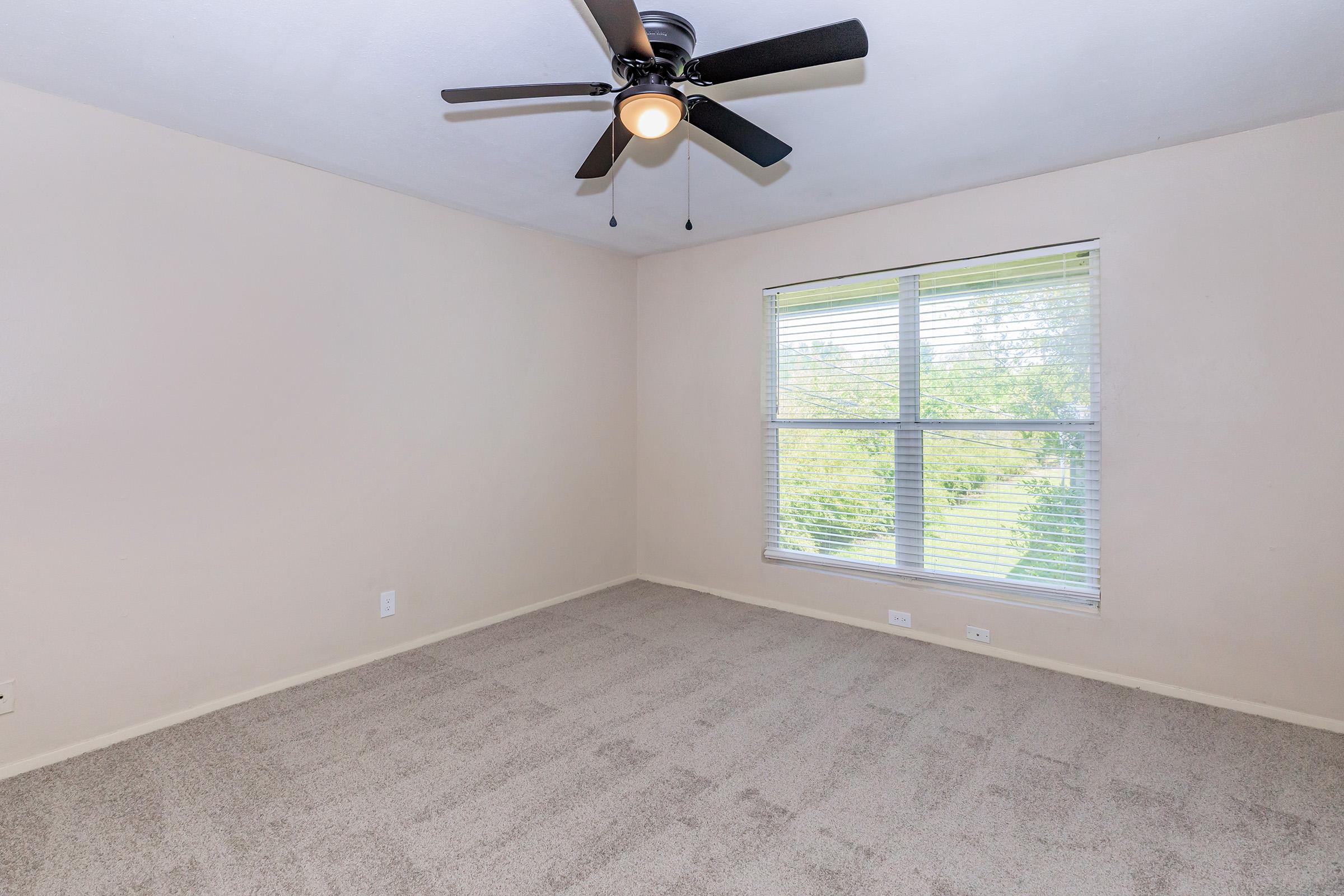 Empty room with light beige walls and carpet, featuring a ceiling fan with four blades. A large window with blinds allows natural light, providing a view of greenery outside. The space appears clean and ready for furnishing.