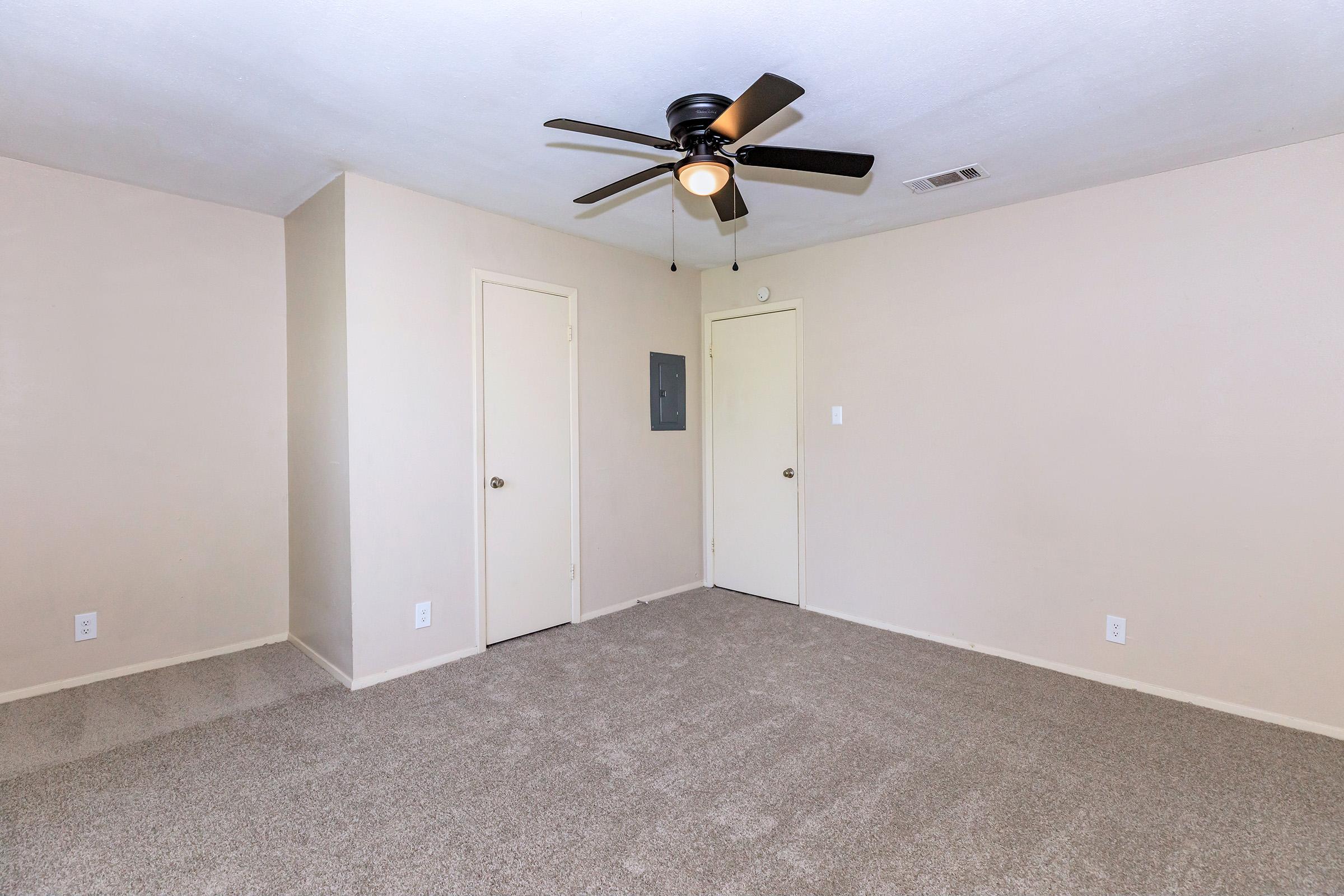 Empty room featuring light beige walls, a light gray carpeted floor, and a ceiling fan with a light. There are two closed doors on the left side, and a gray electrical panel on the wall. The space is well-lit and has a simple, minimalistic design.