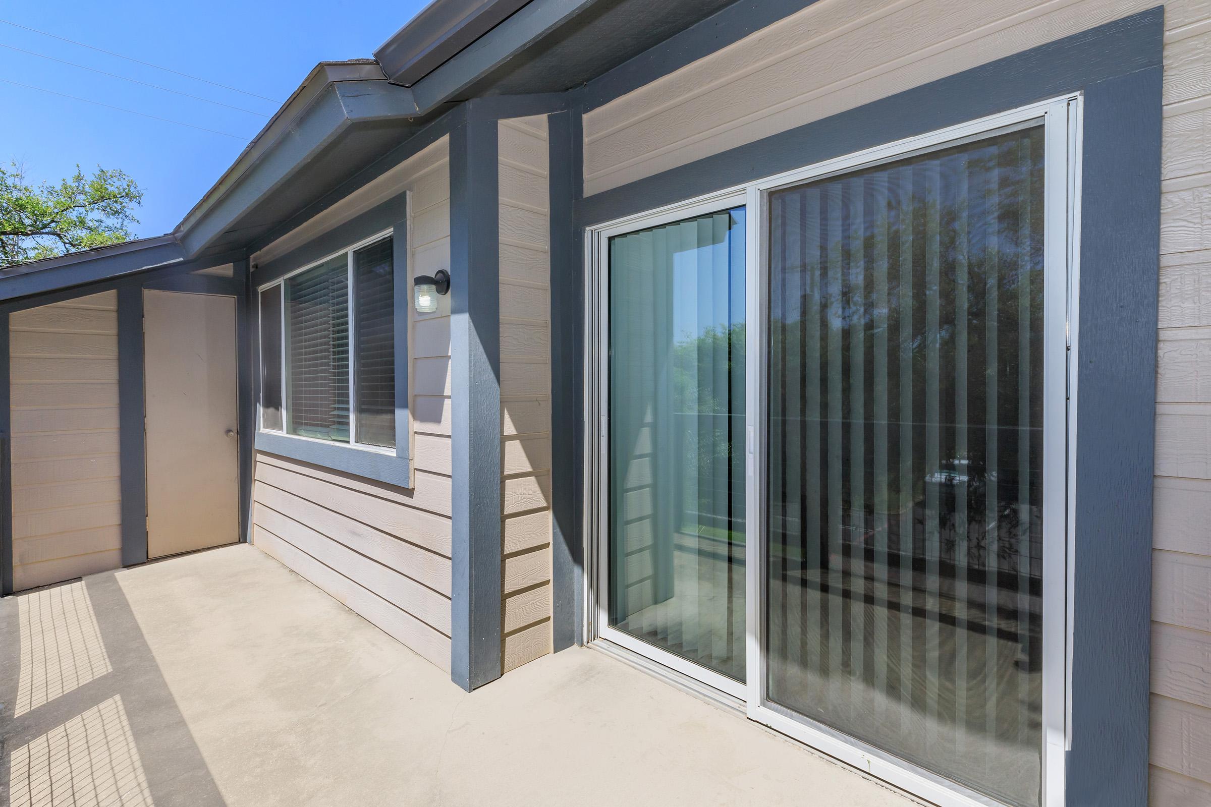 A modern home exterior featuring a sliding glass door with vertical blinds, surrounded by light-colored siding. Natural light enters, illuminating the patio area. The scene is set against a clear blue sky, highlighting a clean and inviting outdoor space.