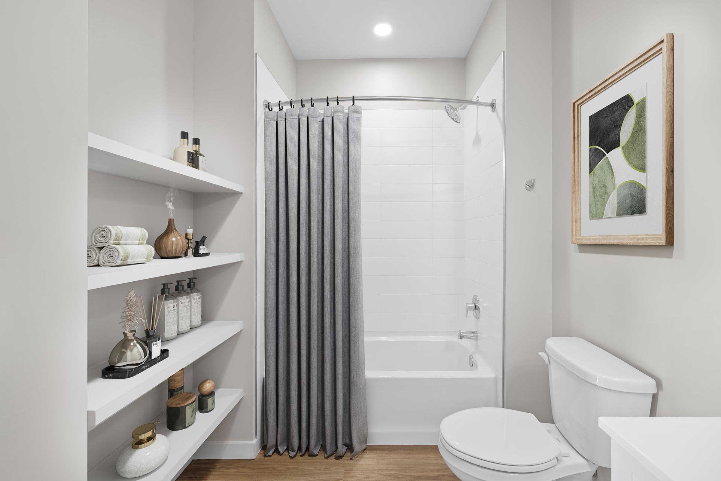 A modern bathroom featuring a bathtub with a shower curtain, white tiled walls, a shelf with neatly arranged toiletries, and decorative elements. There is a framed artwork on the wall, and warm wooden flooring adds a touch of elegance to the space.