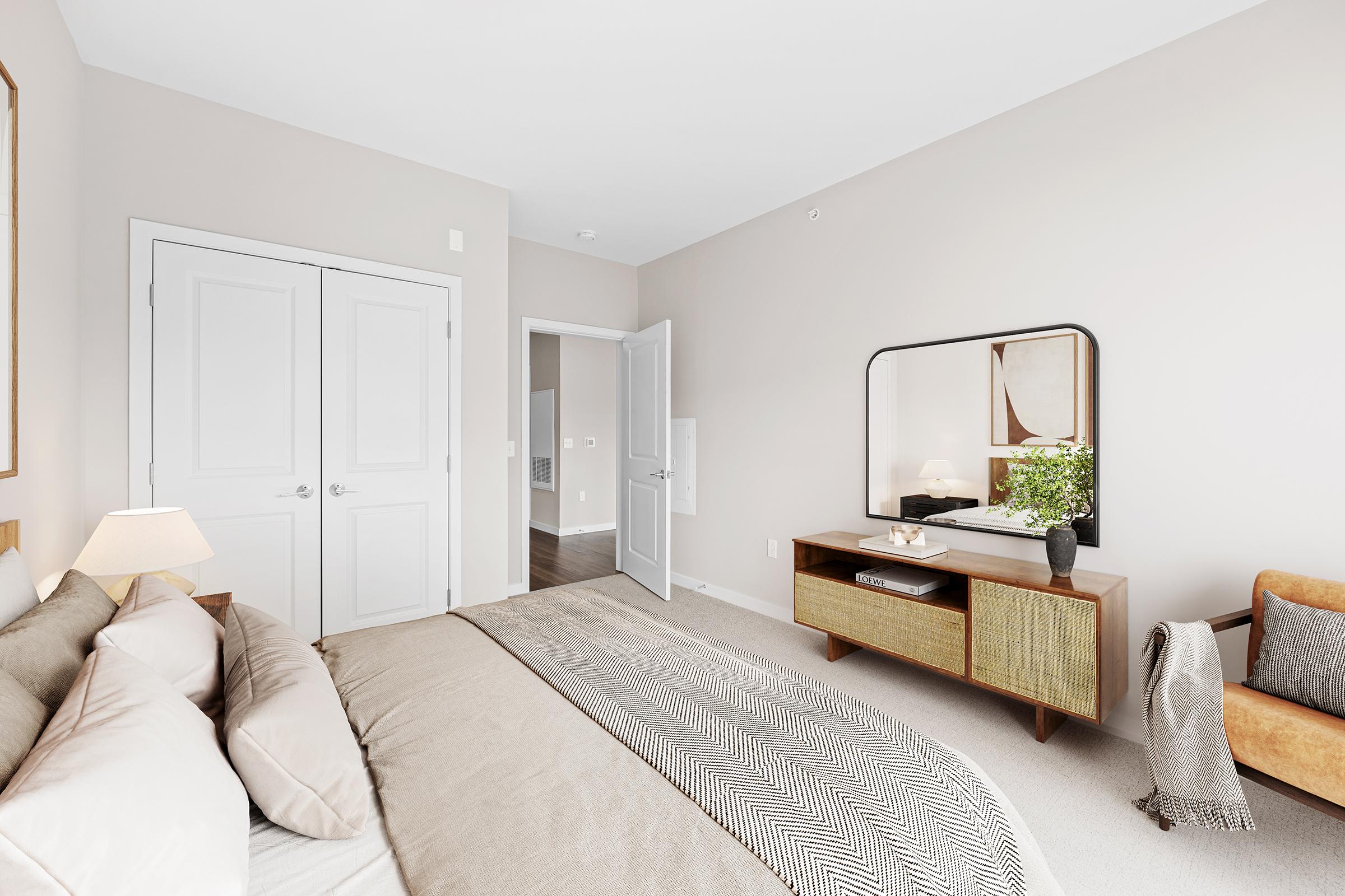 A modern bedroom with a large beige bed adorned with pillows, pastel walls, and a stylish mirror above a dresser. A cozy chair sits in the corner, complemented by a small decorative plant. The room features a door leading to another space and includes a warm, inviting atmosphere.