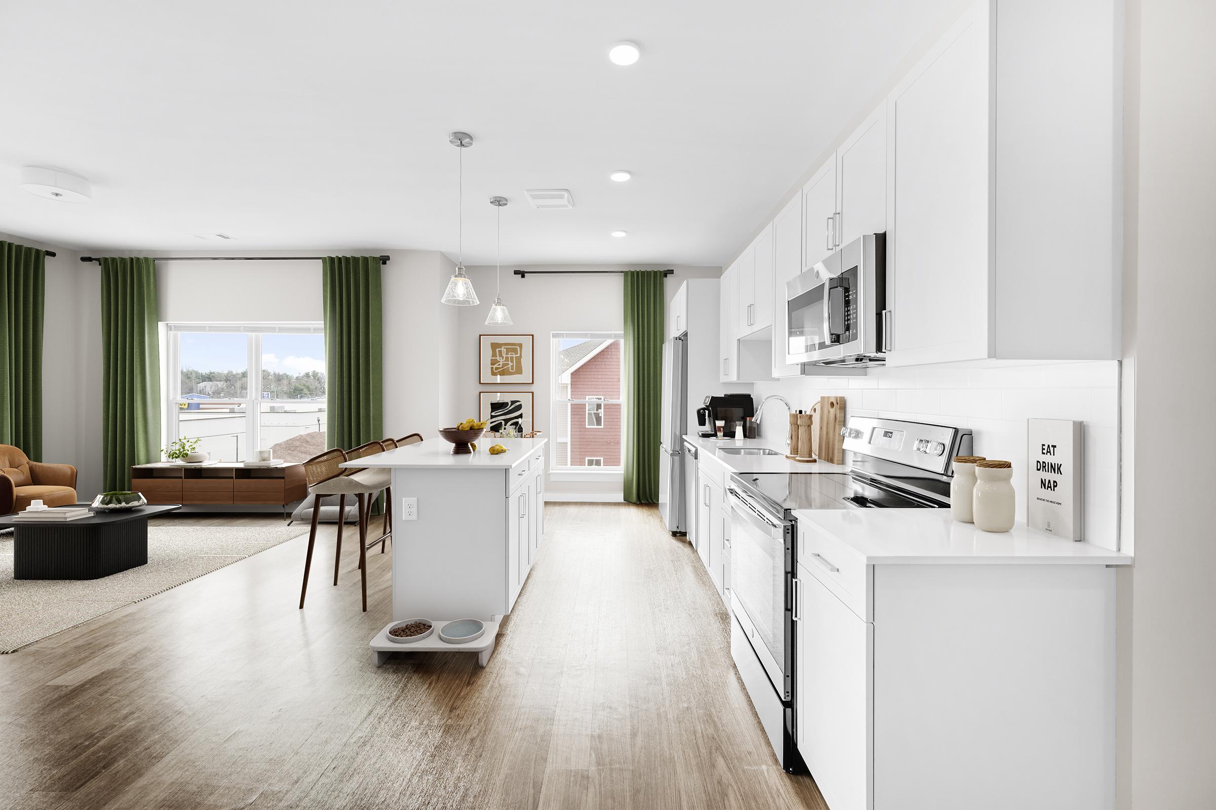 A modern kitchen with white cabinetry and stainless steel appliances, featuring a central island with bar stools. The open layout connects to a living area with green curtains and a cozy seating arrangement. Natural light floods in from large windows, enhancing the fresh and bright ambiance of the space.