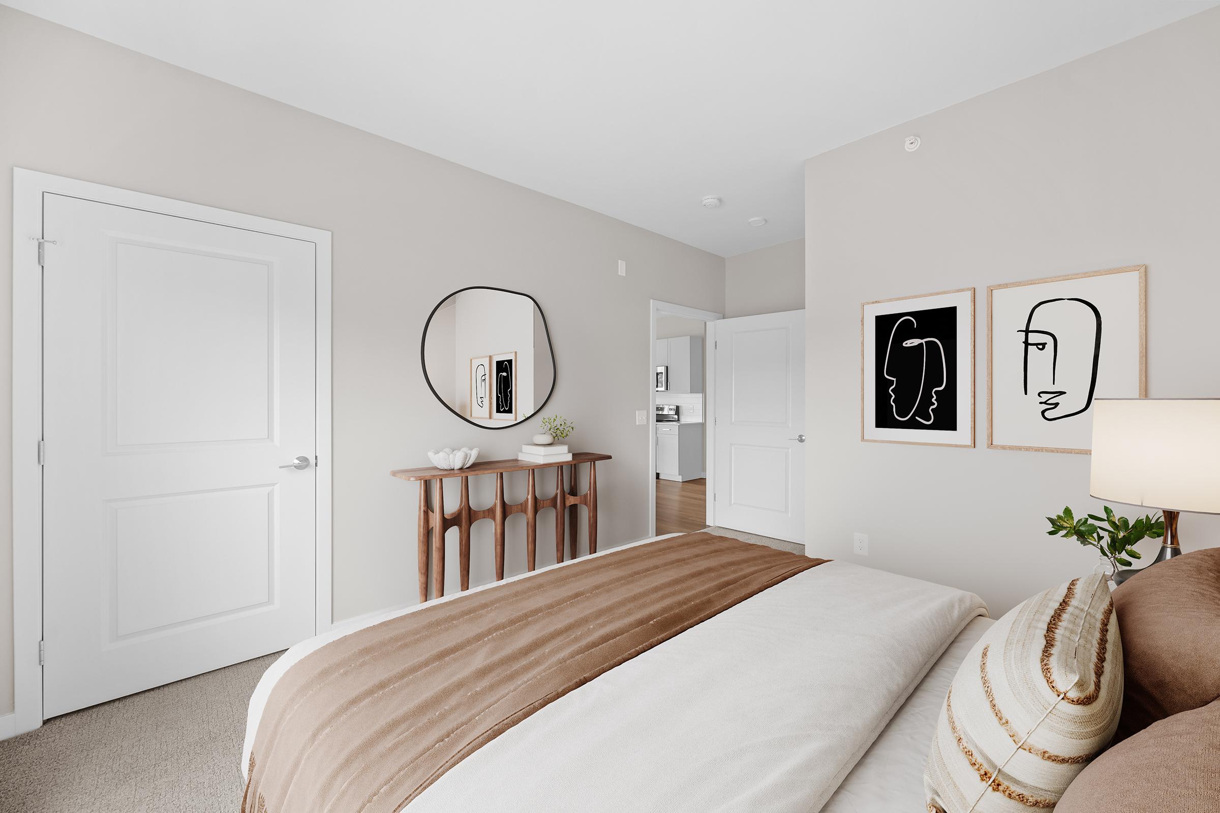 A cozy bedroom featuring a large bed with a brown and white striped blanket. Decor includes a round mirror and two framed abstract line art pieces on the wall. A small wooden console table holds a vase with greenery. The walls are painted in a neutral tone, and a door leads to another room in the background.