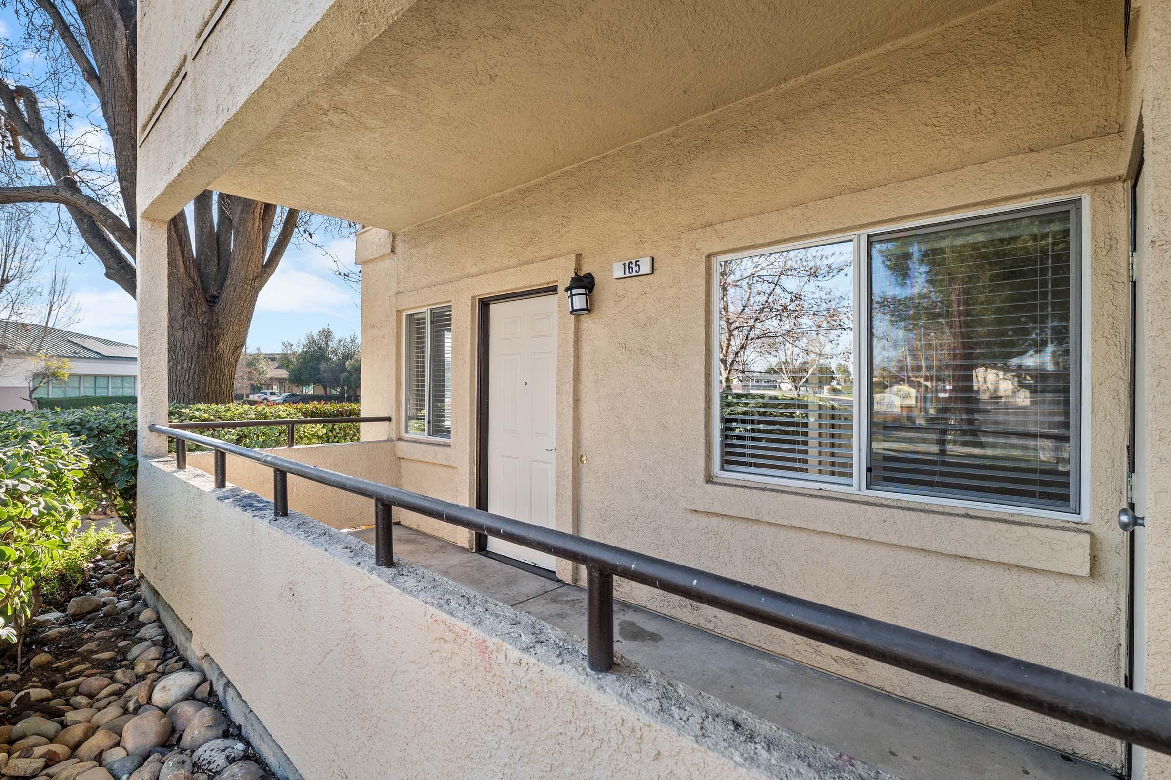 View of a ground-level entrance featuring a white door with a light fixture beside it. The wall is beige, and there are large windows with blinds. A railing runs along a small porch area, with a landscaped garden visible in the foreground, including rocks and greenery. Bright daylight illuminates the scene.