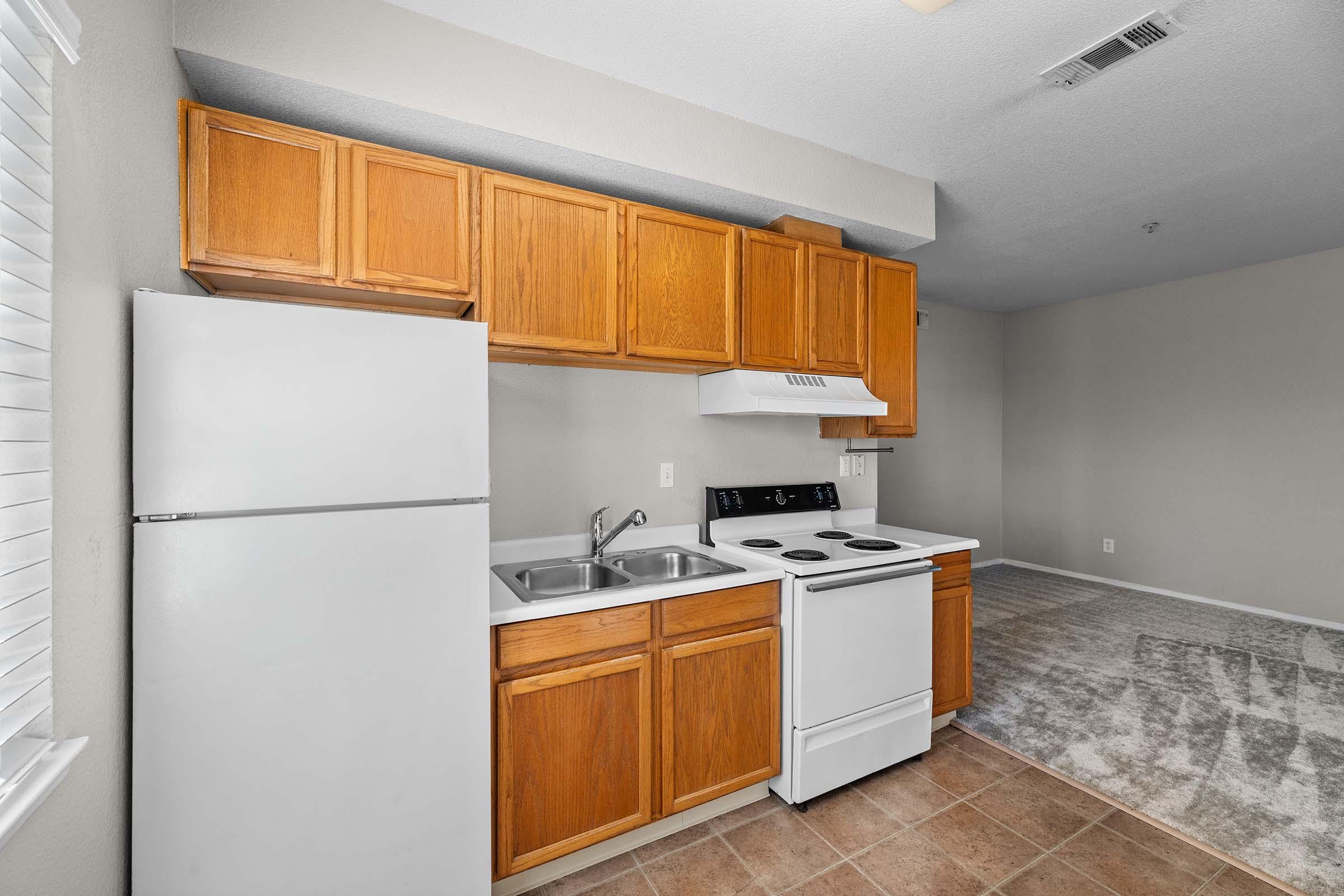 A small kitchen space featuring wooden cabinetry, a white refrigerator, a stove with an oven, and a sink. The floor is tiled, and there is a light-colored carpet in the adjoining area. Natural light enters through a window with blinds.