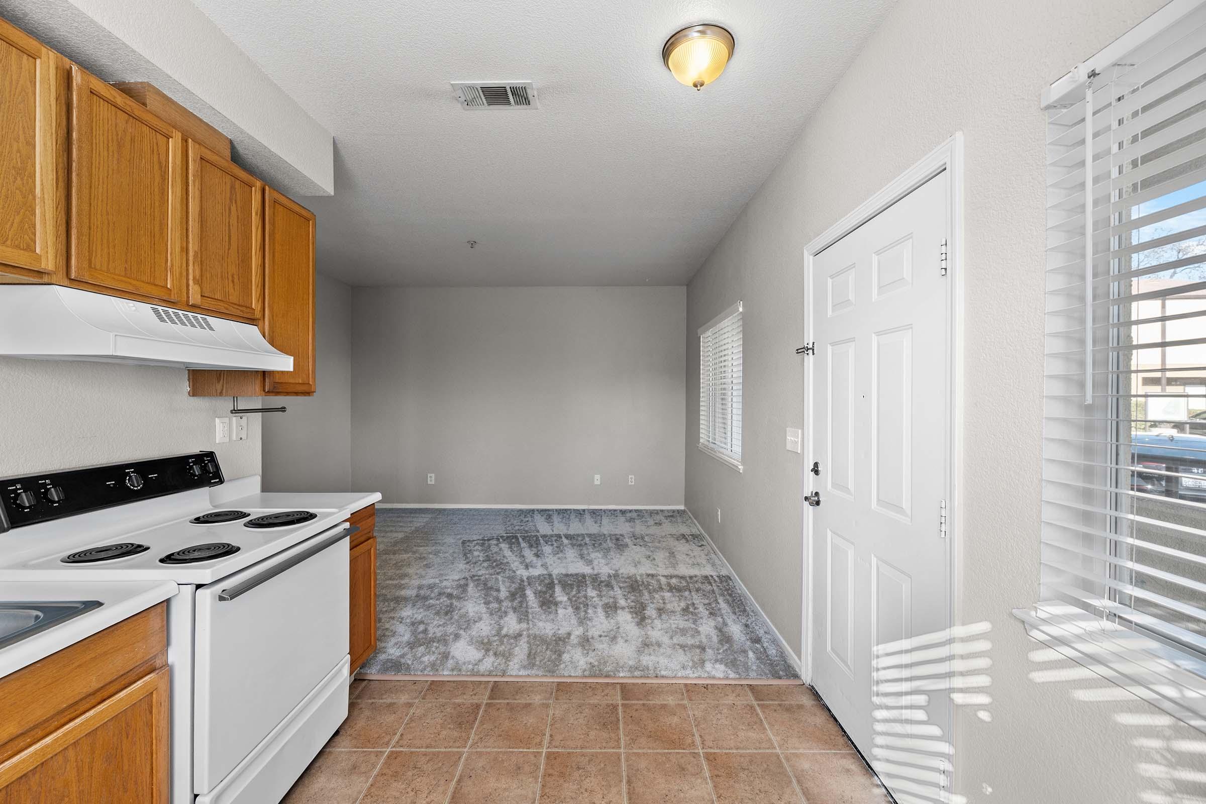 A kitchen area featuring wooden cabinets, a white stove, and a tile floor. Adjacent to the kitchen is an empty living space with gray carpet and walls, a window with blinds, and a front door leading outside. Natural light is coming through the window, creating a bright atmosphere.