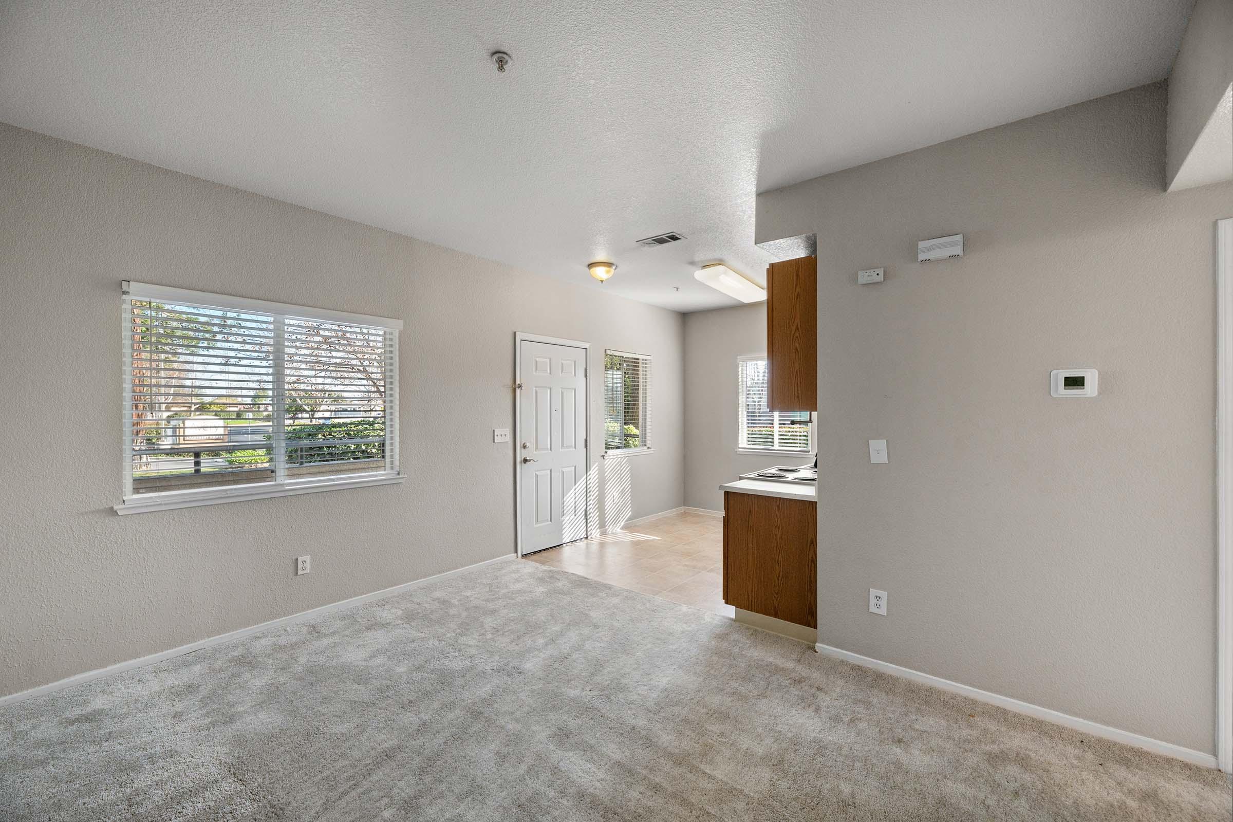 A light-filled room featuring beige carpet and freshly painted walls. There is a door leading outside, a window with blinds, and a small kitchen area with wooden cabinets. The space appears open and inviting, perfect for a cozy living area or an apartment living space.