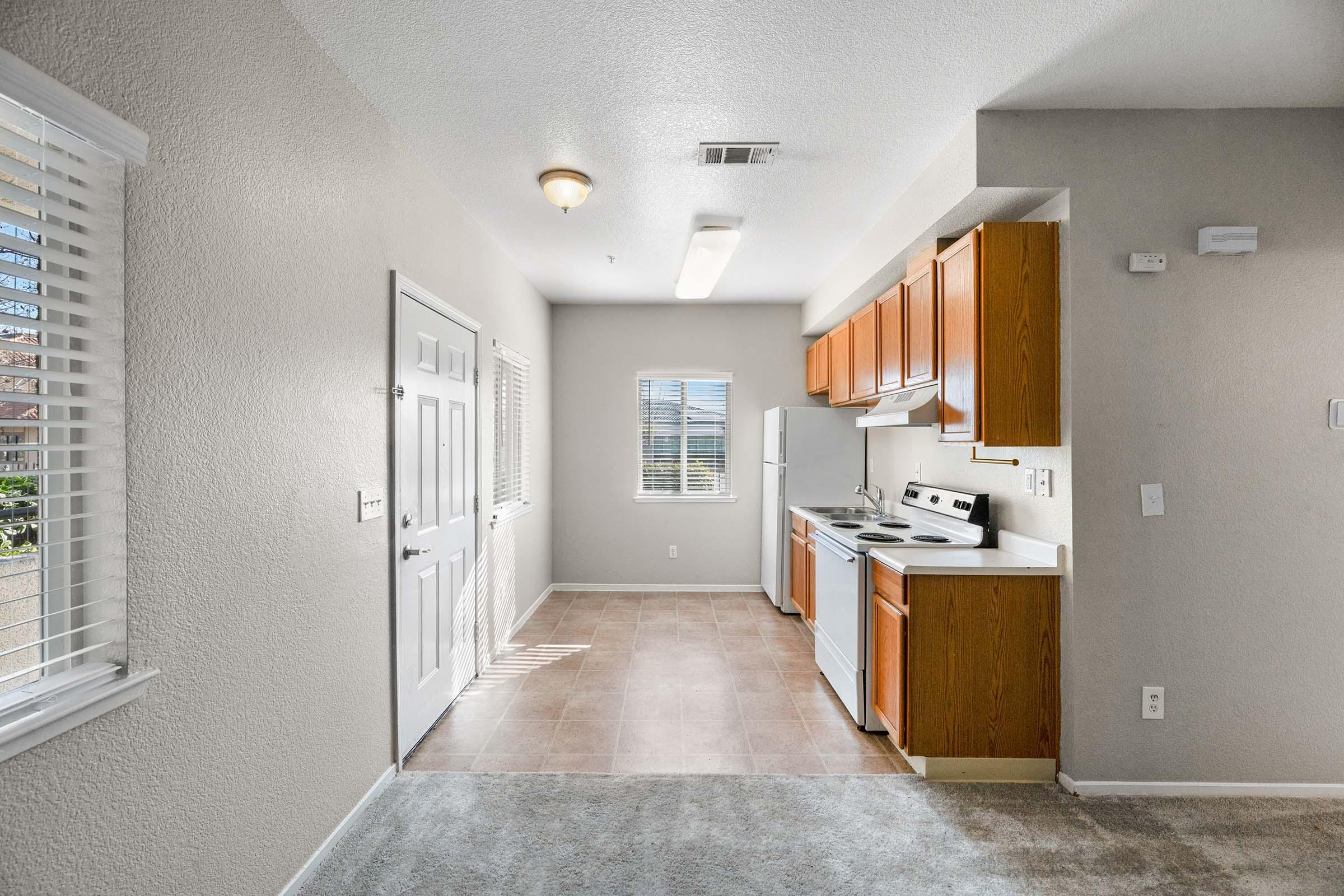 Interior view of a bright and spacious kitchen and dining area. Features include light-colored walls, a white stove, and wooden cabinets. Natural light streams in through a window, illuminating the tiled floor and carpeted area. A door leads to the outside, enhancing the open feel of the space.
