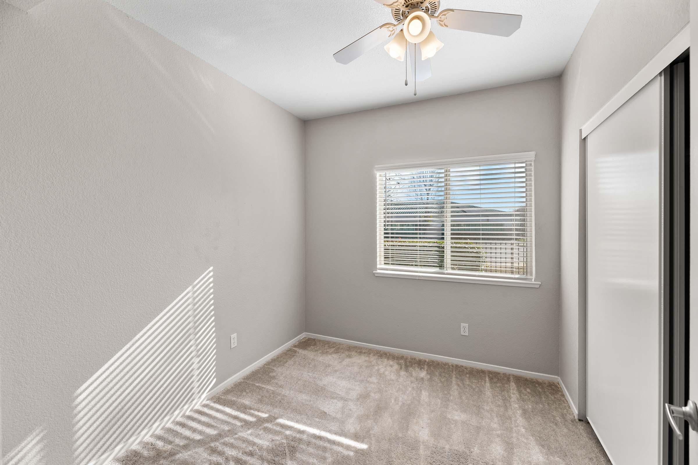 Empty room with light gray walls and a light carpet. A ceiling fan is mounted in the center, and there is a window with blinds that allows natural light to enter. Shadows from the blinds form stripes on the carpeted floor. The room has a clean and minimalist appearance.