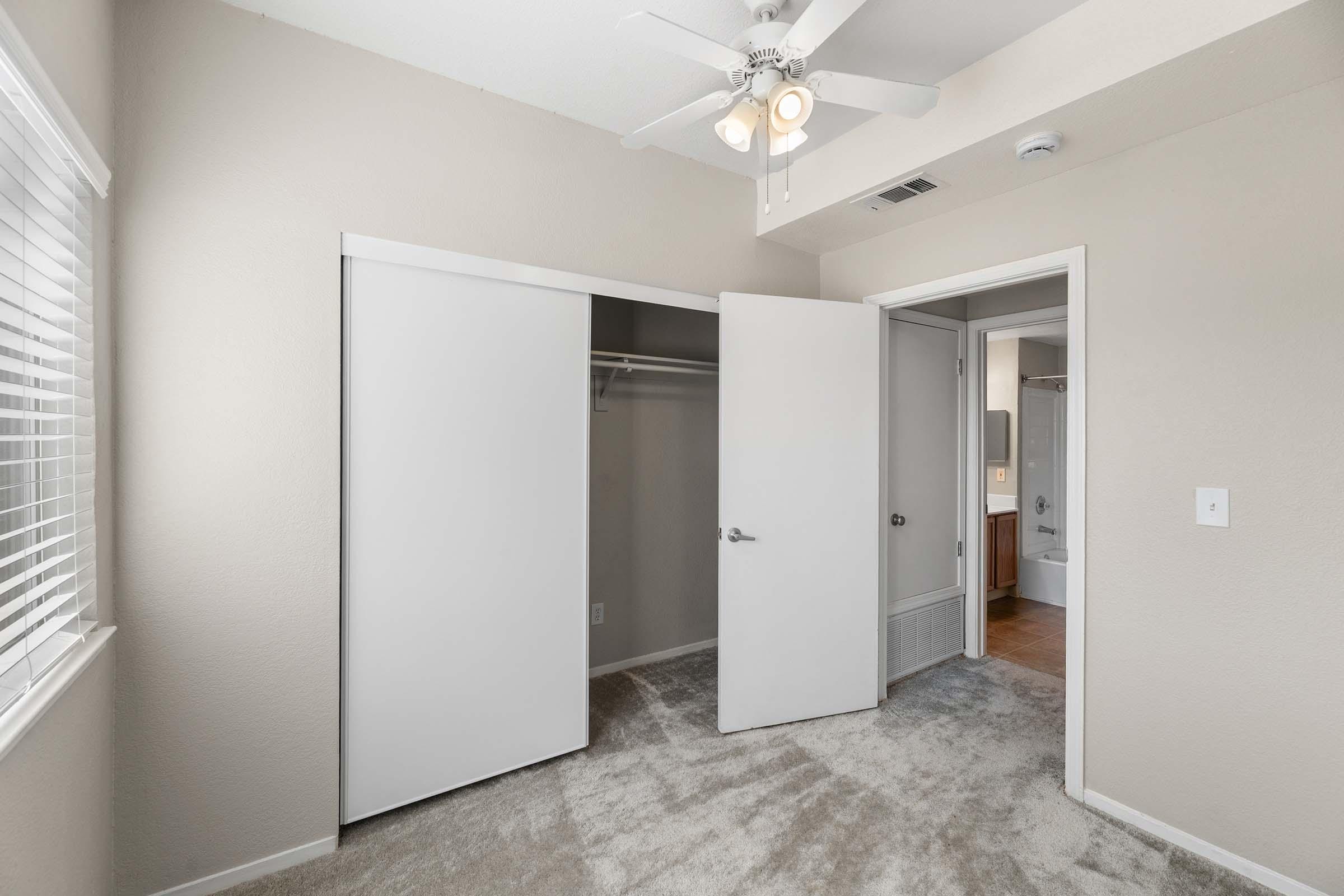A well-lit, empty room with beige walls and plush carpet. It features a ceiling fan, a large closet with sliding doors, and an adjoining bathroom visible through an open door. Windows allow natural light, and the space conveys a clean, minimalist aesthetic.