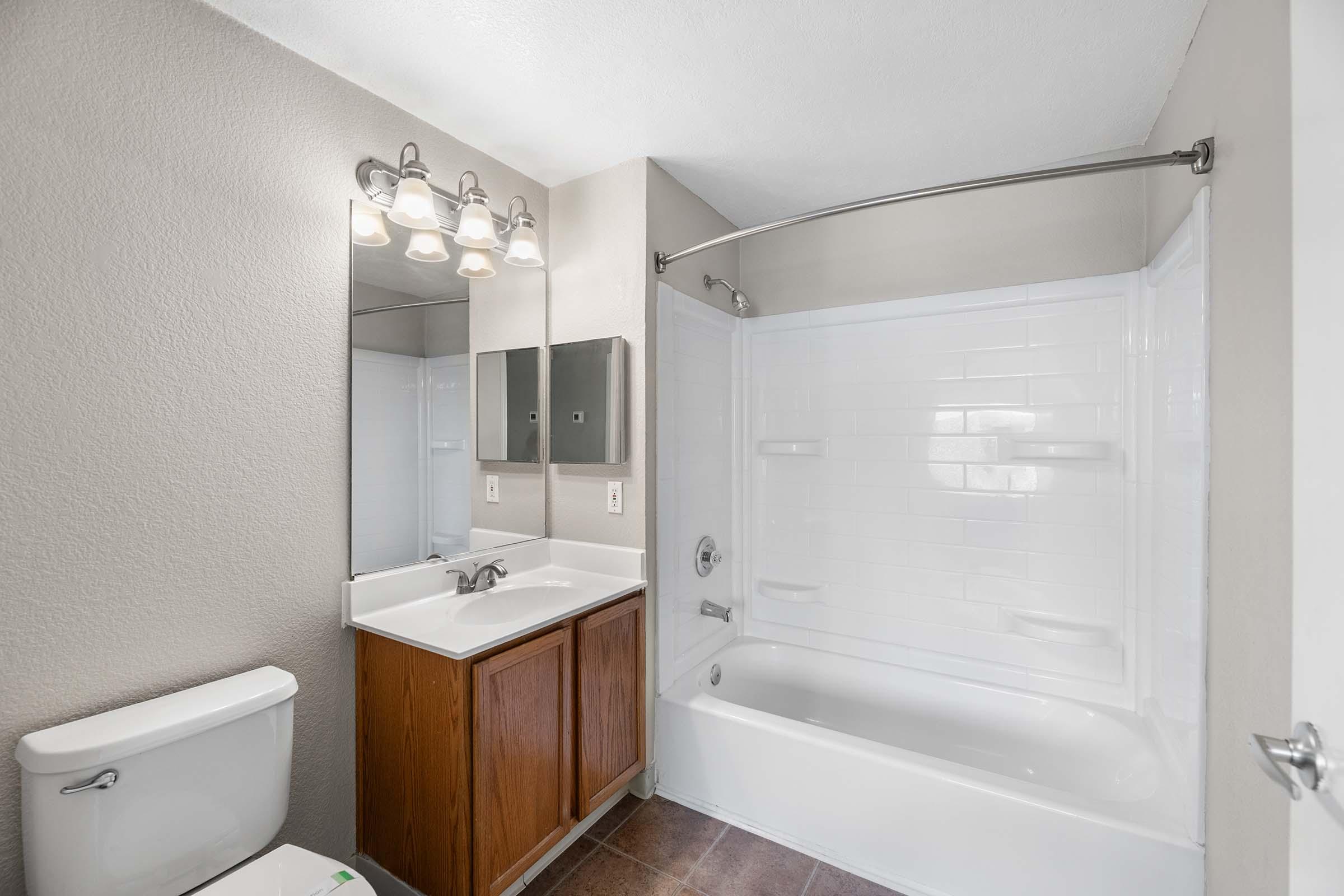 A clean, modern bathroom featuring a white bathtub with shower, a sink under a mirror, an overhead light fixture, and wooden cabinetry. The walls are painted in a light color, and there is a toilet in the corner, creating a spacious and bright environment.