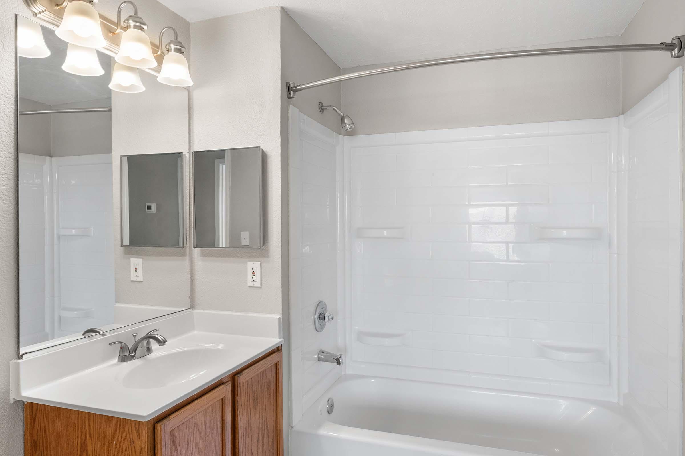 A modern bathroom featuring a white bathtub and shower enclosure, a single sink with wooden cabinetry, and two small mirrored cabinets above the sink. Soft overhead lighting illuminates the space, which has neutral-colored walls.