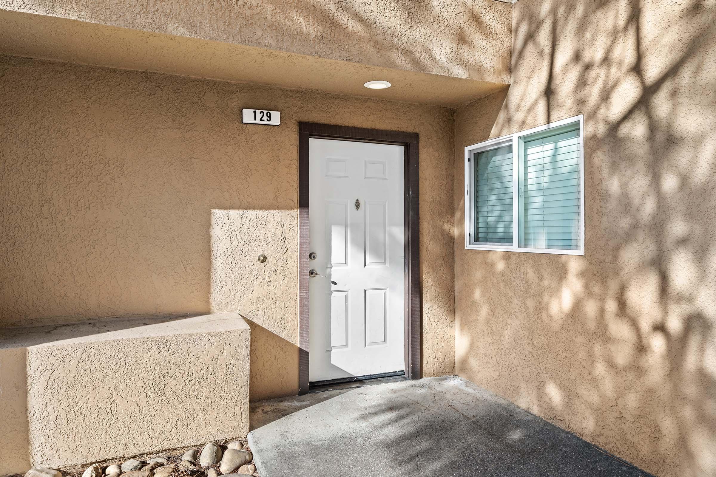 A front entrance with a white door and a small window next to it, set against a textured beige wall. The door has a number "129" above it, and there is a small, irregularly shaped ledge beside it. Shadows from surrounding foliage are visible on the wall.