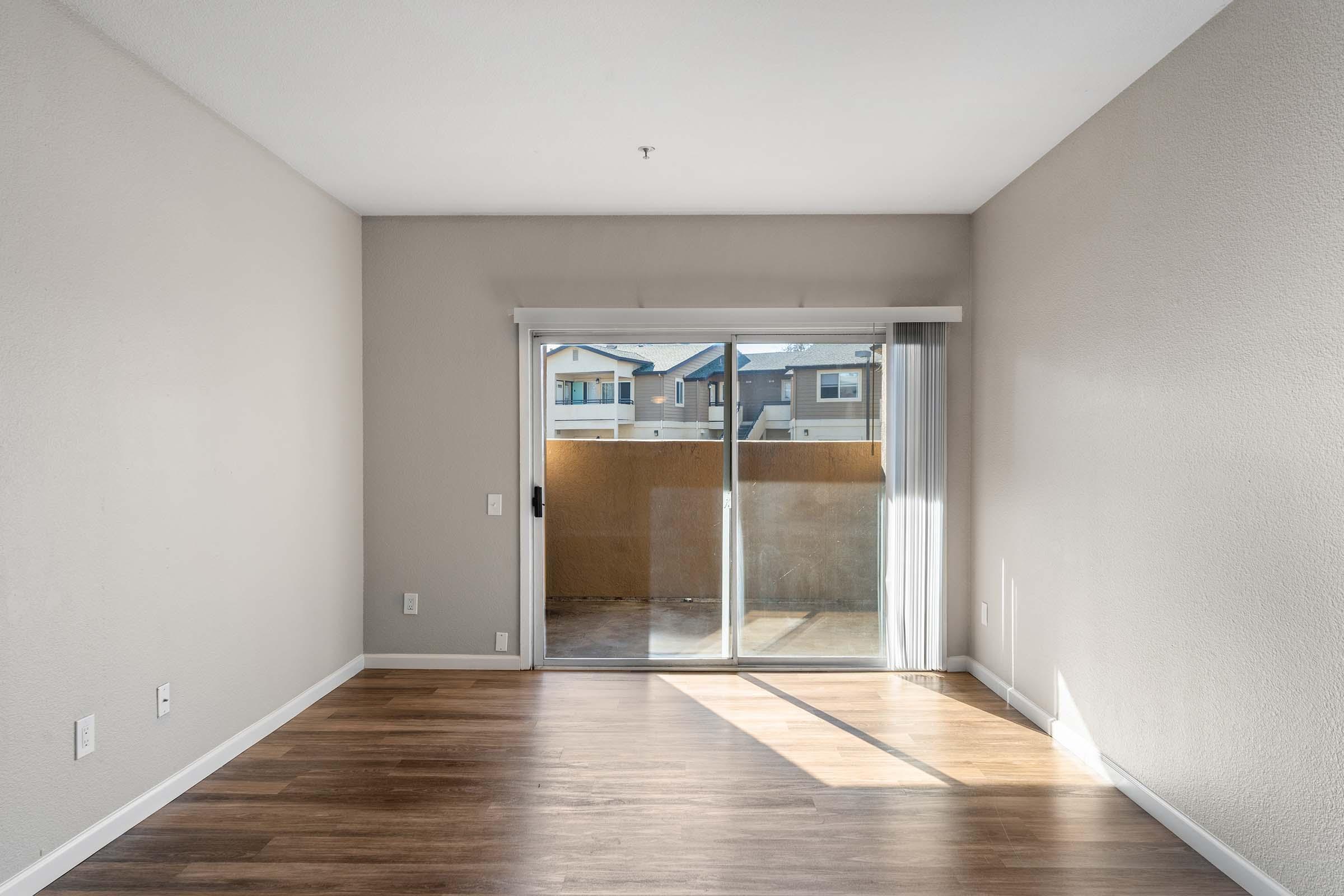 A spacious, unfurnished living room with light-colored walls and wood laminate flooring. A large sliding glass door leads to a small balcony, allowing natural light to fill the room. The opposite wall is bare with electrical outlets visible. Bright and airy atmosphere, perfect for personalizing.