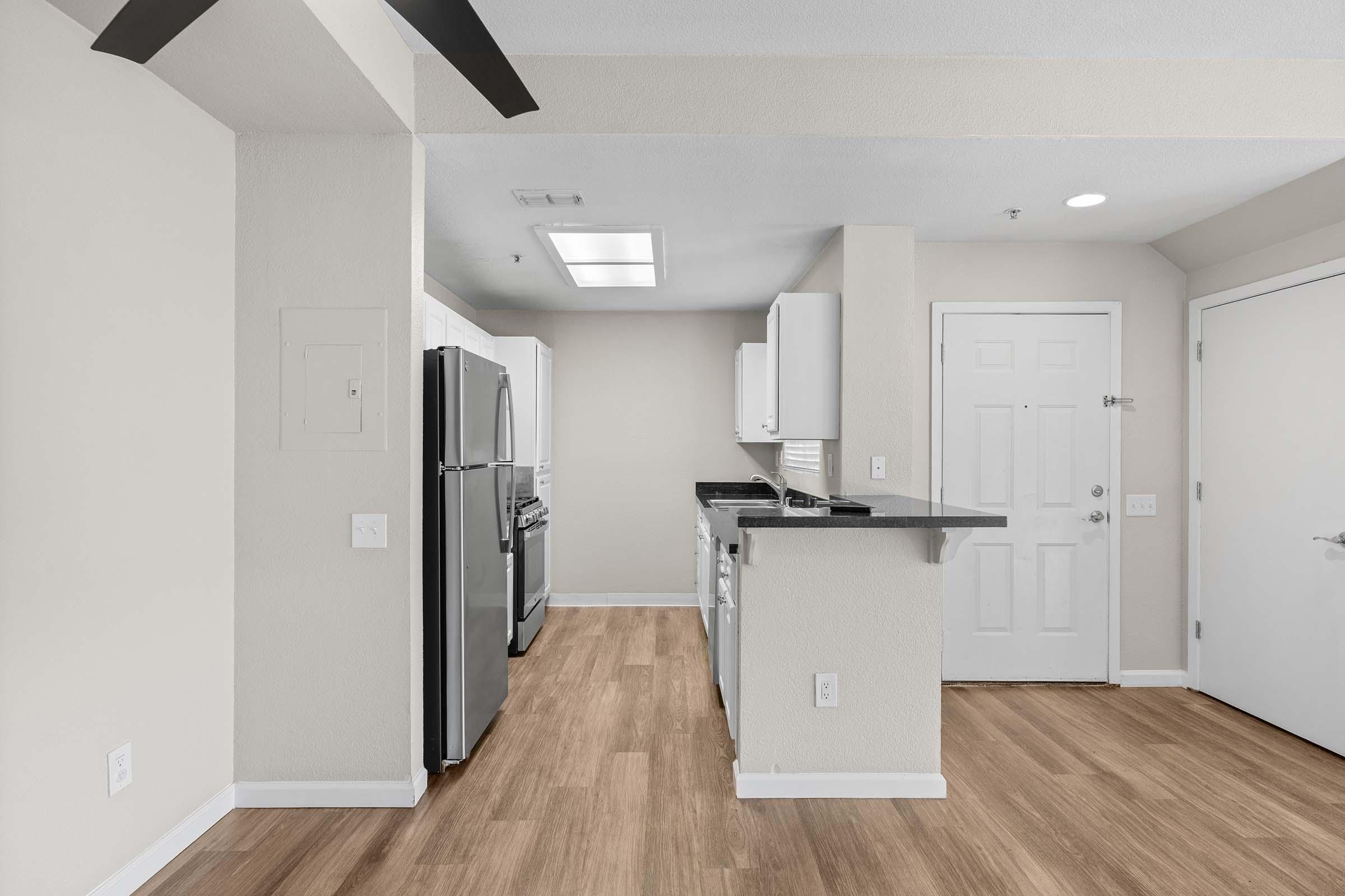 A modern kitchen featuring light-colored cabinets, stainless steel appliances, and a spacious layout with a wooden floor. The entrance door is visible, along with a door to another room. Natural light is coming from a ceiling fixture and potential windows, enhancing the room's bright ambiance.