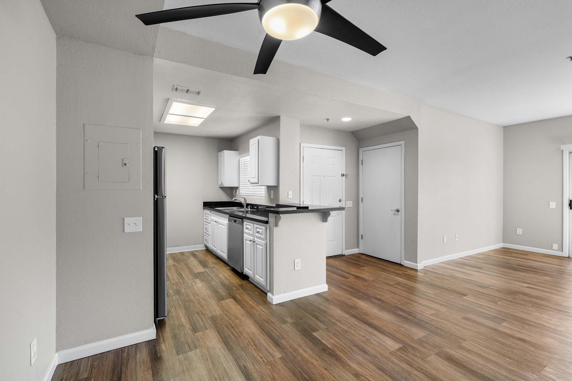 Modern kitchen and living area featuring wood-like flooring, a ceiling fan, white cabinets, and stainless steel appliances. The space is well-lit with a combination of natural light and ceiling fixtures, showcasing a minimalist design with neutral walls and an open layout.
