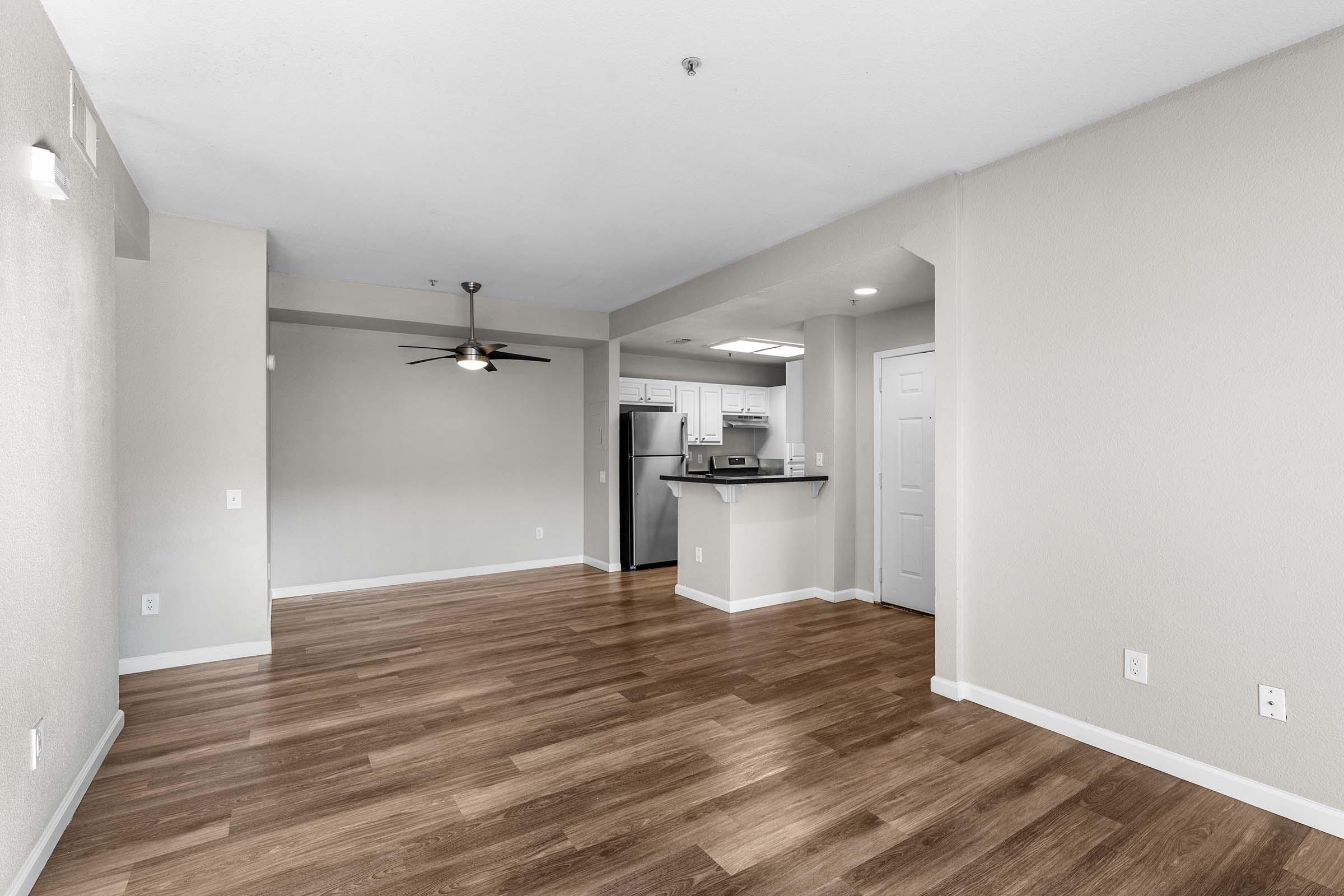 Empty living room with hardwood flooring, a ceiling fan, and light-colored walls. A kitchen area with a bar counter is visible in the background, featuring stainless steel appliances. Natural light fills the space, creating a bright and open atmosphere.