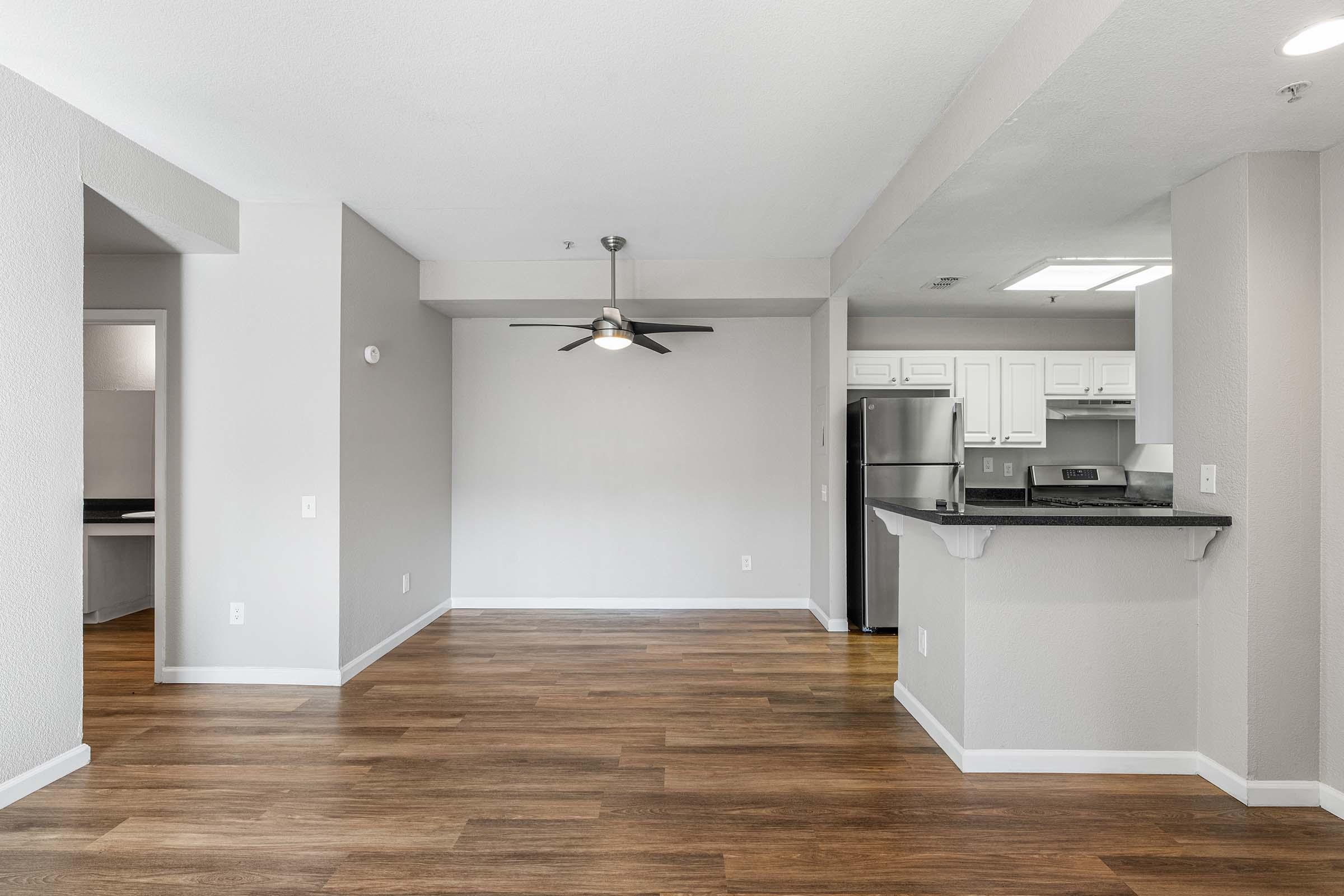 A modern, open-concept living space featuring light gray walls, a ceiling fan, and hardwood-style flooring. The kitchen is visible with stainless steel appliances and white cabinetry. The layout includes a bar area, and natural light fills the room, creating a spacious and inviting atmosphere.