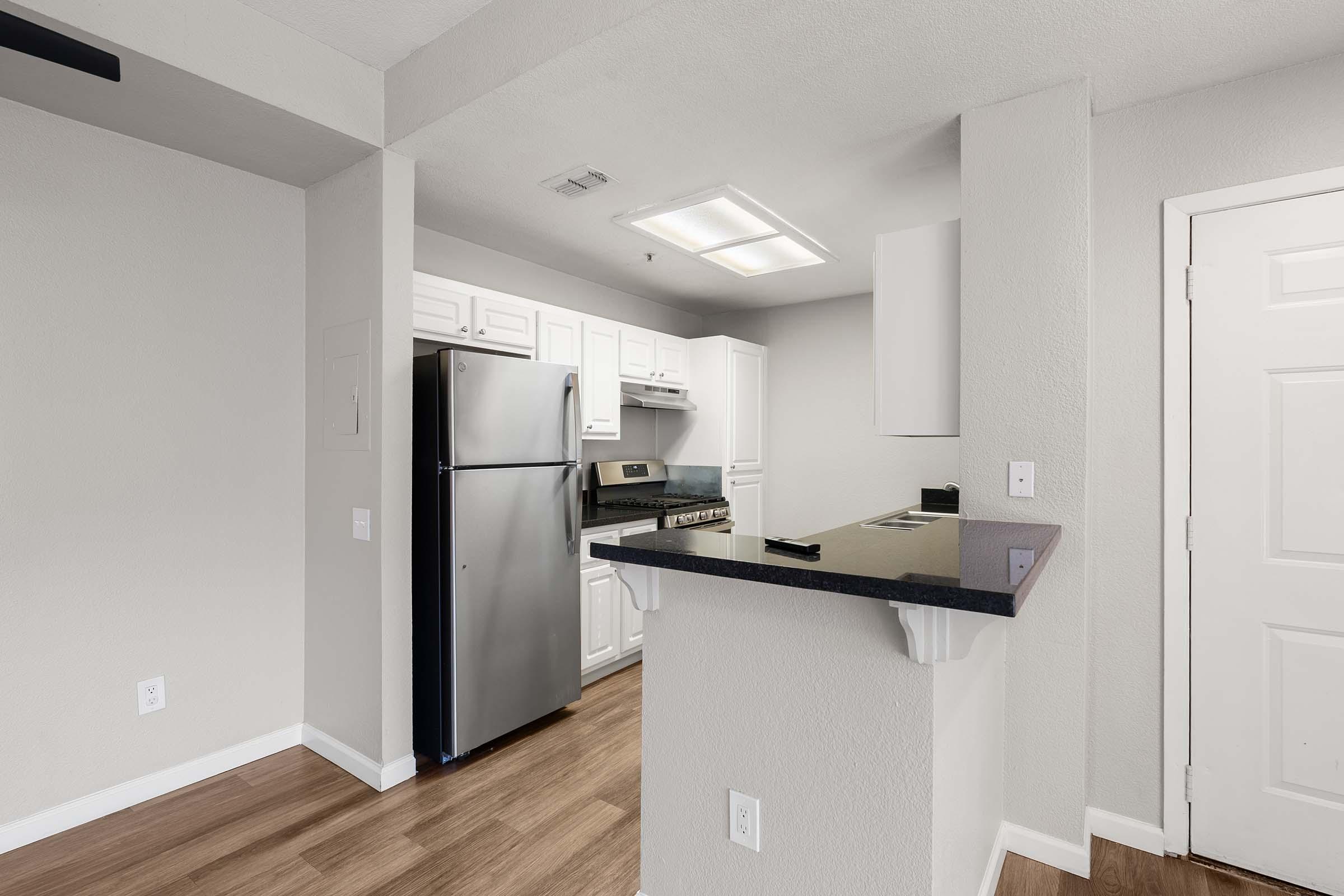 Modern kitchen with white cabinets, stainless steel refrigerator, and stove. The space features wooden flooring, a granite countertop, and an open layout leading to a door on the right. Bright overhead lighting illuminates the room, creating a clean and inviting atmosphere.