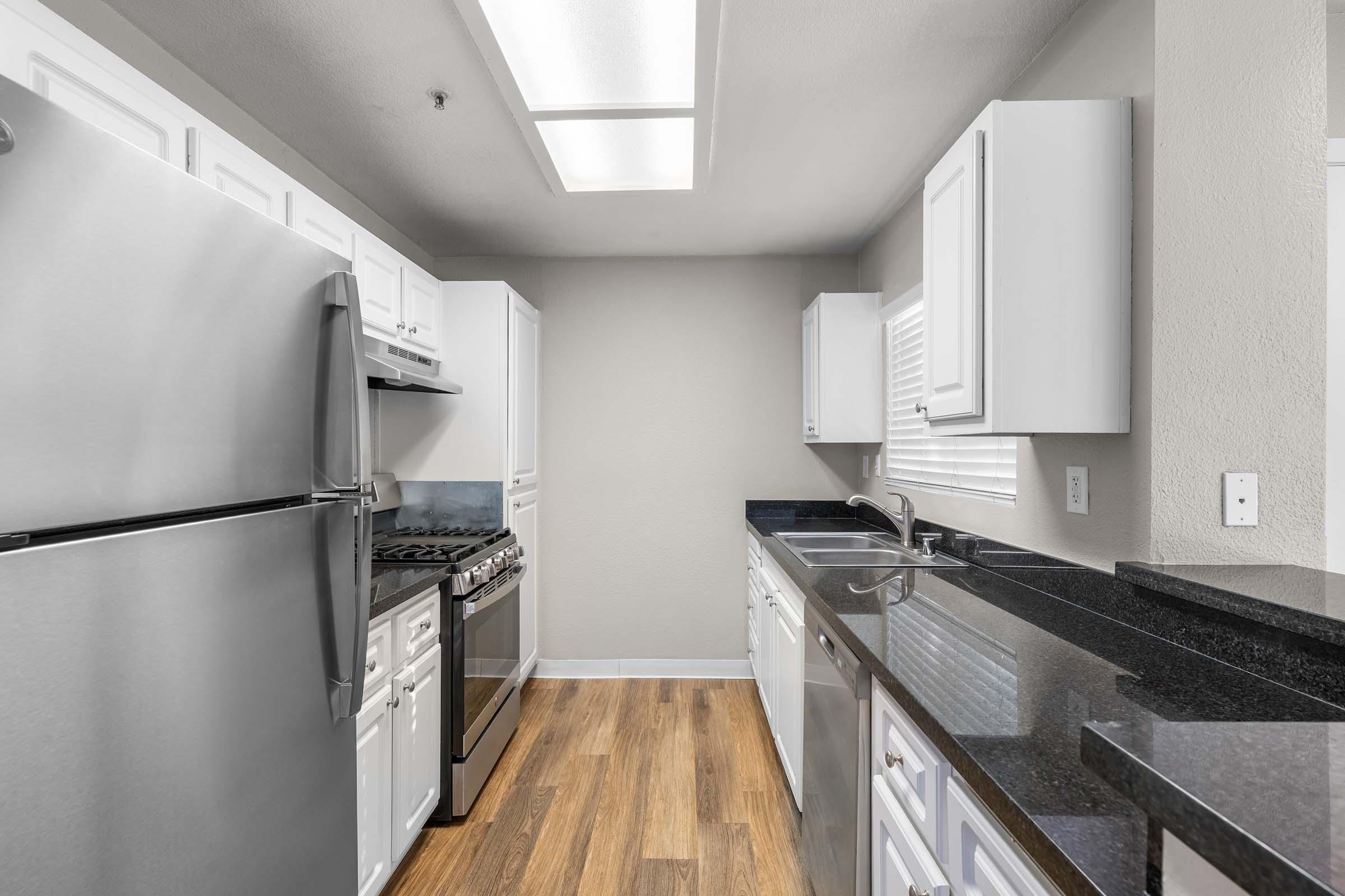 A modern kitchen featuring white cabinetry, stainless steel appliances, including a refrigerator and oven, and a black granite countertop. The space has a light wood floor and is illuminated by natural light from a window. The design is clean and contemporary, emphasizing functionality.